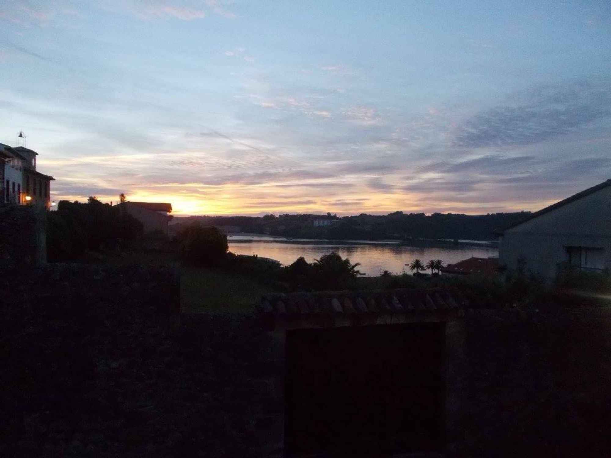 A serene view at sunrise over a river, featuring silhouettes of buildings and lush greenery along the water's edge. Soft colors in the sky blend from orange to blue, reflecting on the water's surface.