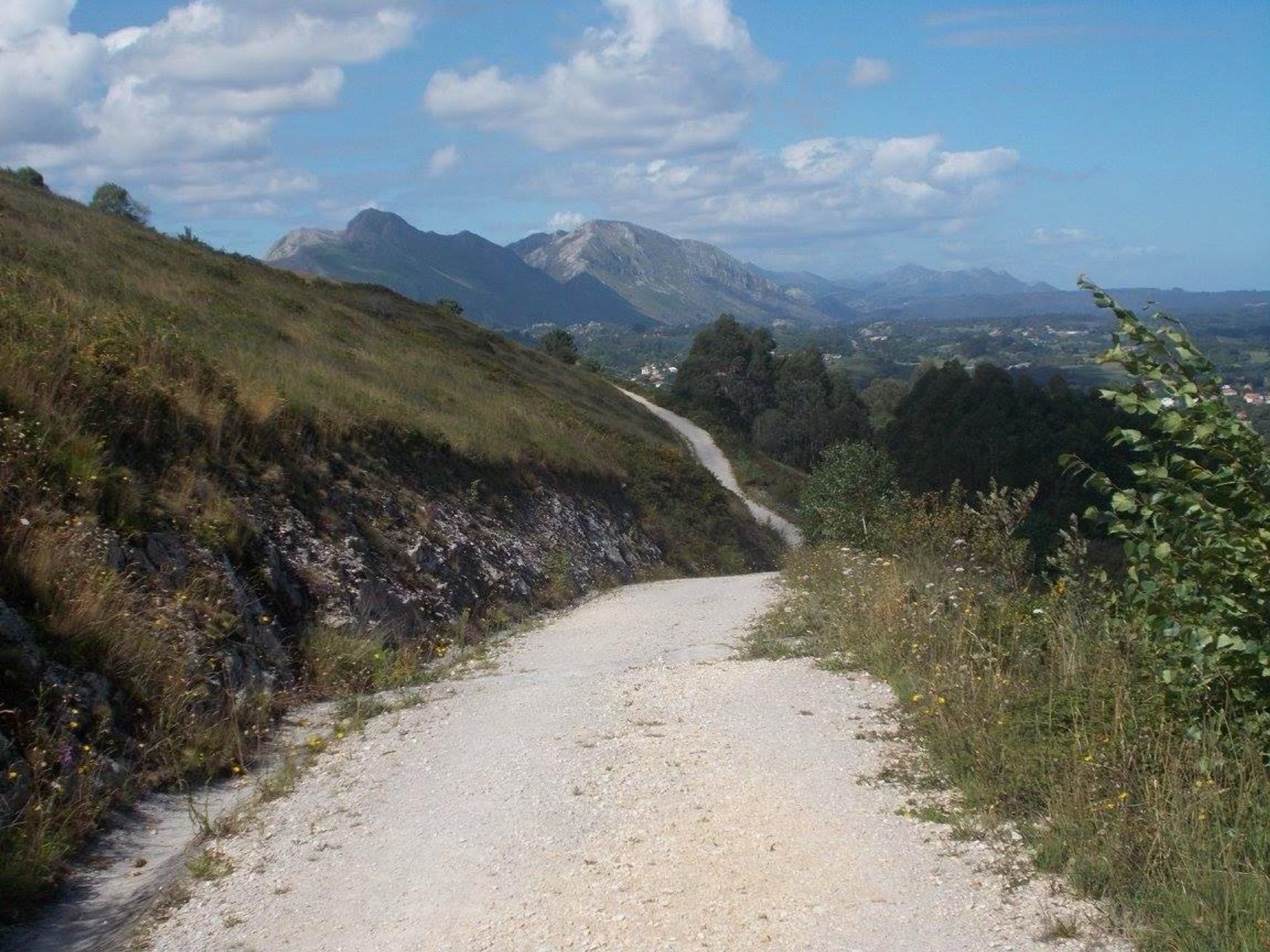 A winding gravel path leads through grassy terrain, with mountains visible in the background under a partly cloudy sky.