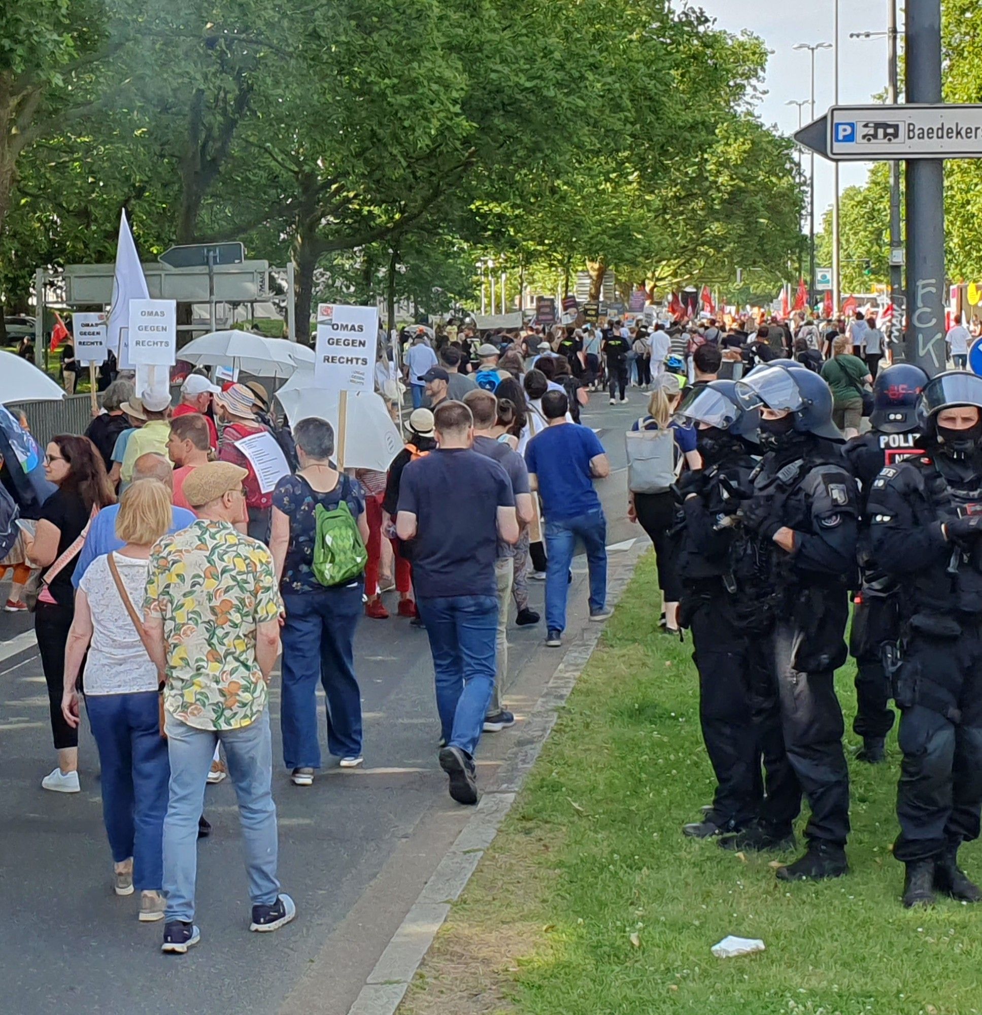 Entlang der Demoroute gehen auf der linken Seite Demonstrantinnen eine Straße entlang. Auf der rechten Seite stehen auf einem Grünstreifen behelmte Einsatzkräfte der Polizei in voller Schutzausrüstung.