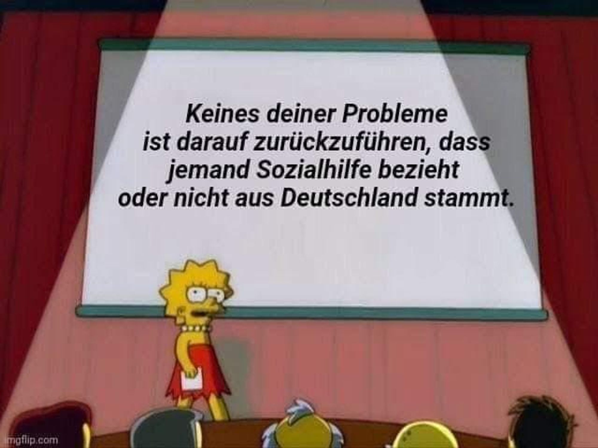 Lisa Simpson steht auf einer Bühne vor Publikum, von dem man nur halbe Hinterköpfe sieht. Hinter ihr ist eine große Leinwand auf welcher steht: Keines Deiner Probleme ist darauf zurückzuführen, dass jemand Sozialhilfe bezieht oder nicht aus Deutschland stammt.