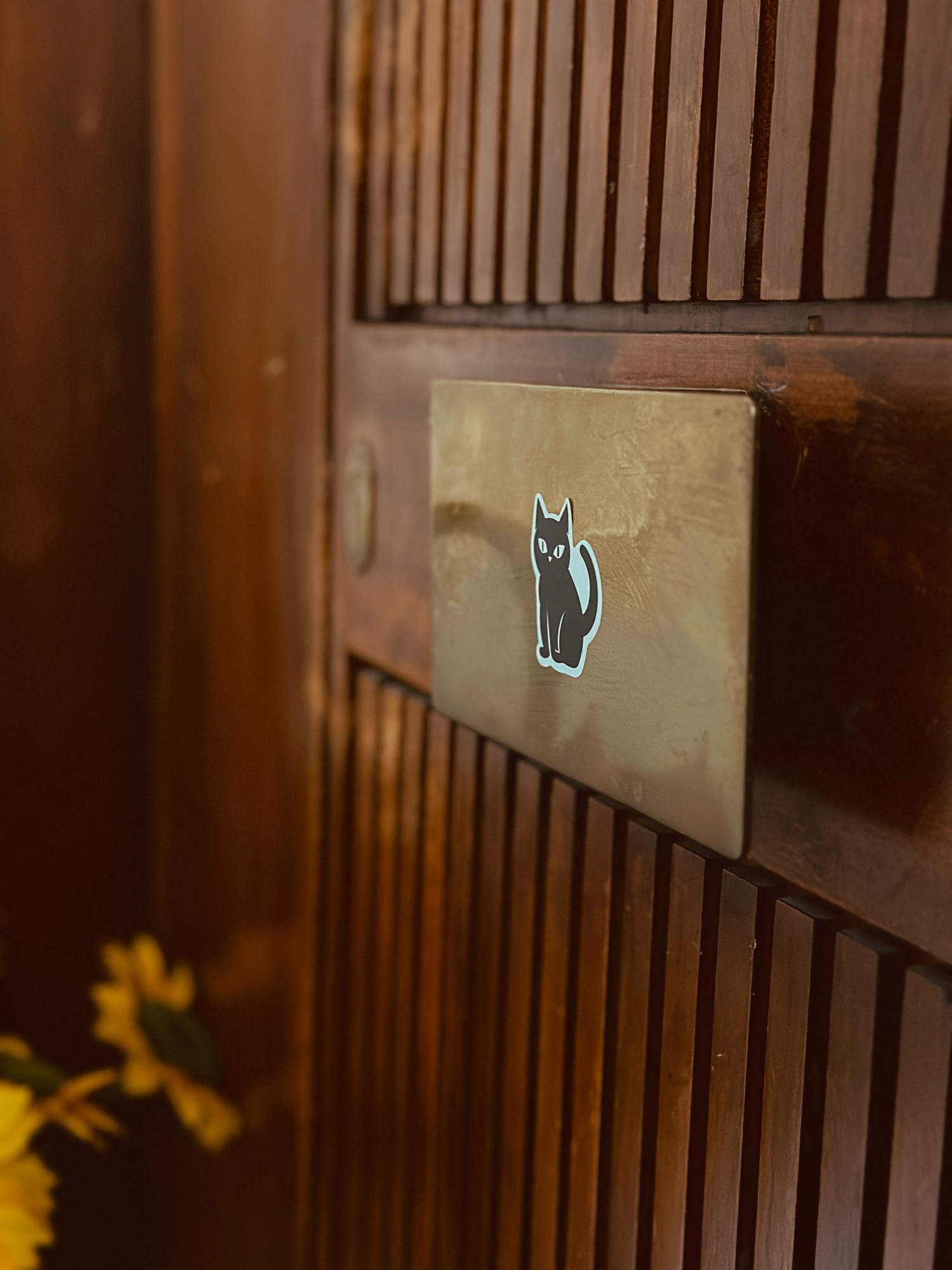 A dark, wooden door with a brass handle that has a small, black and white sticker of a cute cat on it. In the lower left corner, a bunch of sunflowers. 