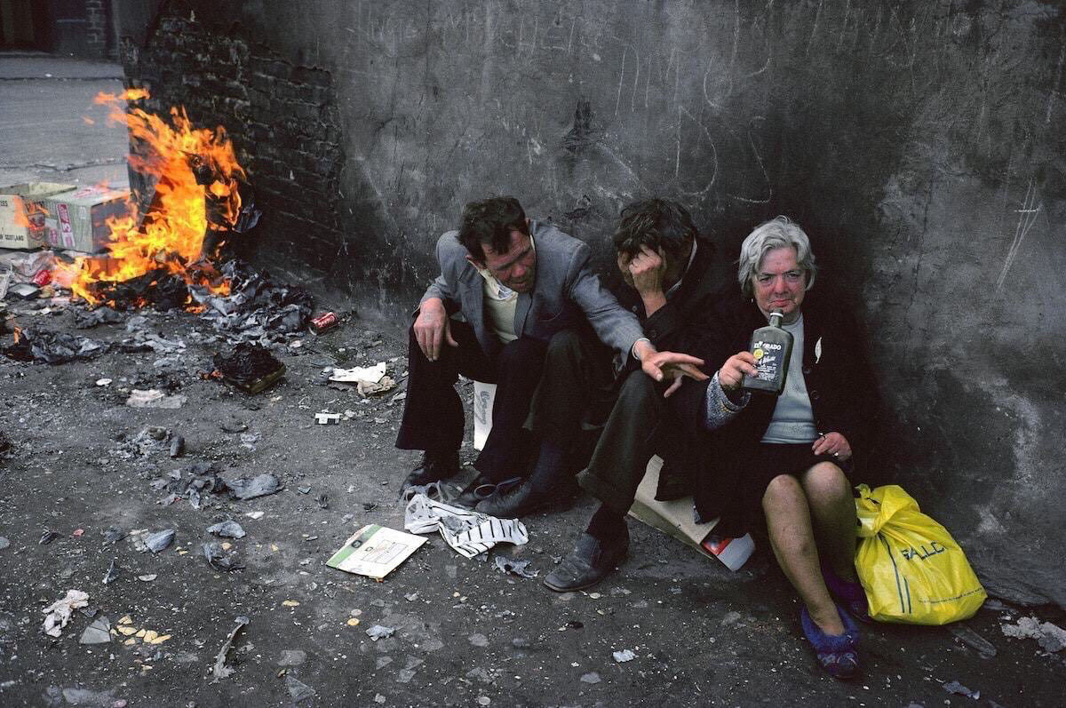 Colour photo of three people sitting on the ground drinking with a fire burning in the background 