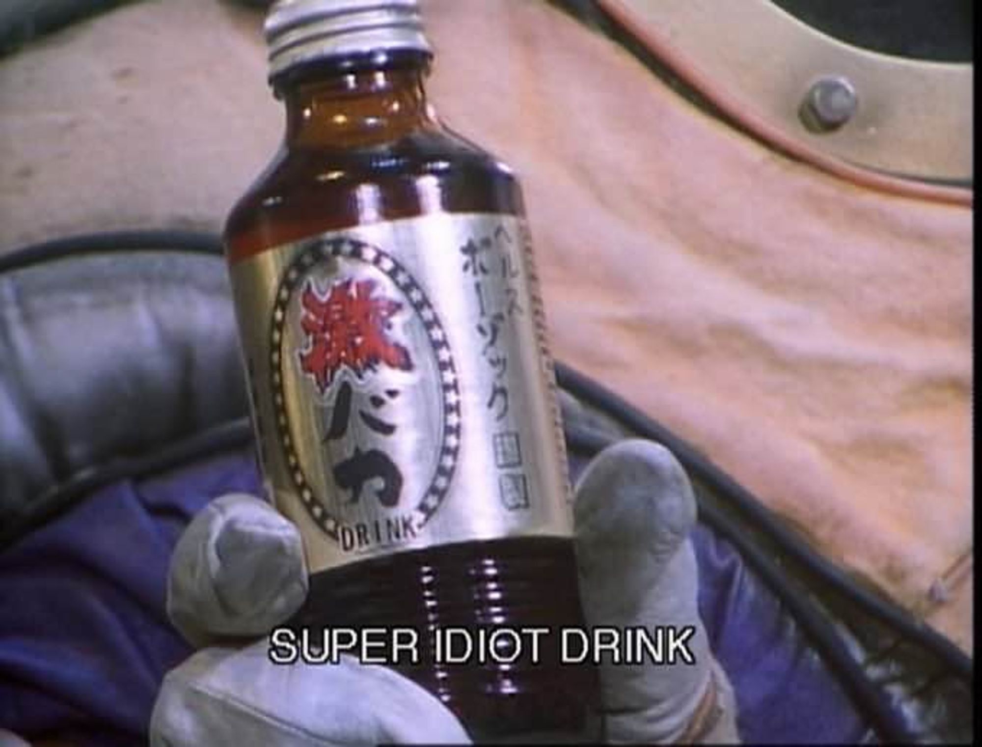 A brown glass bottle with a silver lable with Japanese writing on it. The subtitles explain the label reads "Super idiot drink"