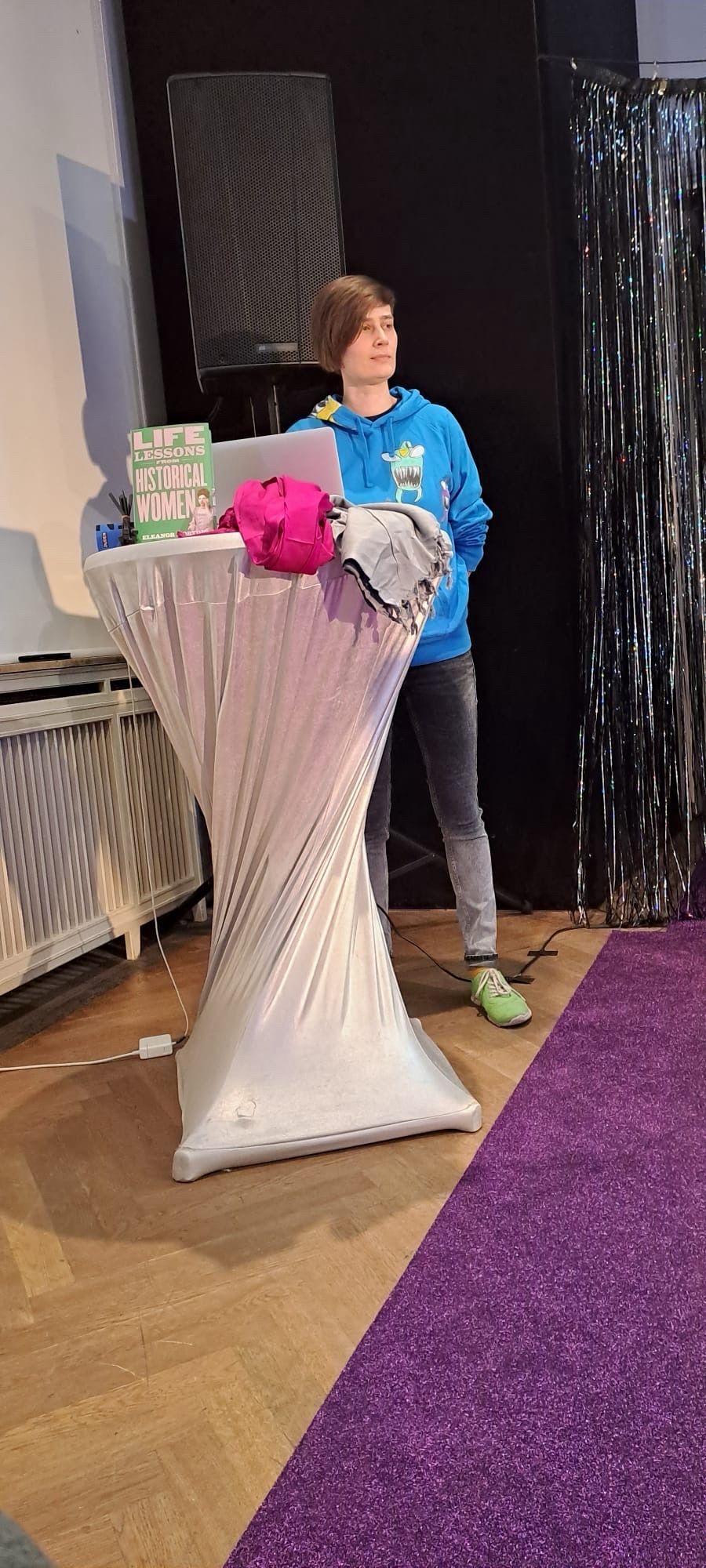 Me standing behind a high table in a silver cover. On the table are 2 scarves, a book by Eleanor Morton and my laptop. Me and the table are right next to the edge of a purple glittery carpet that marks the stage.