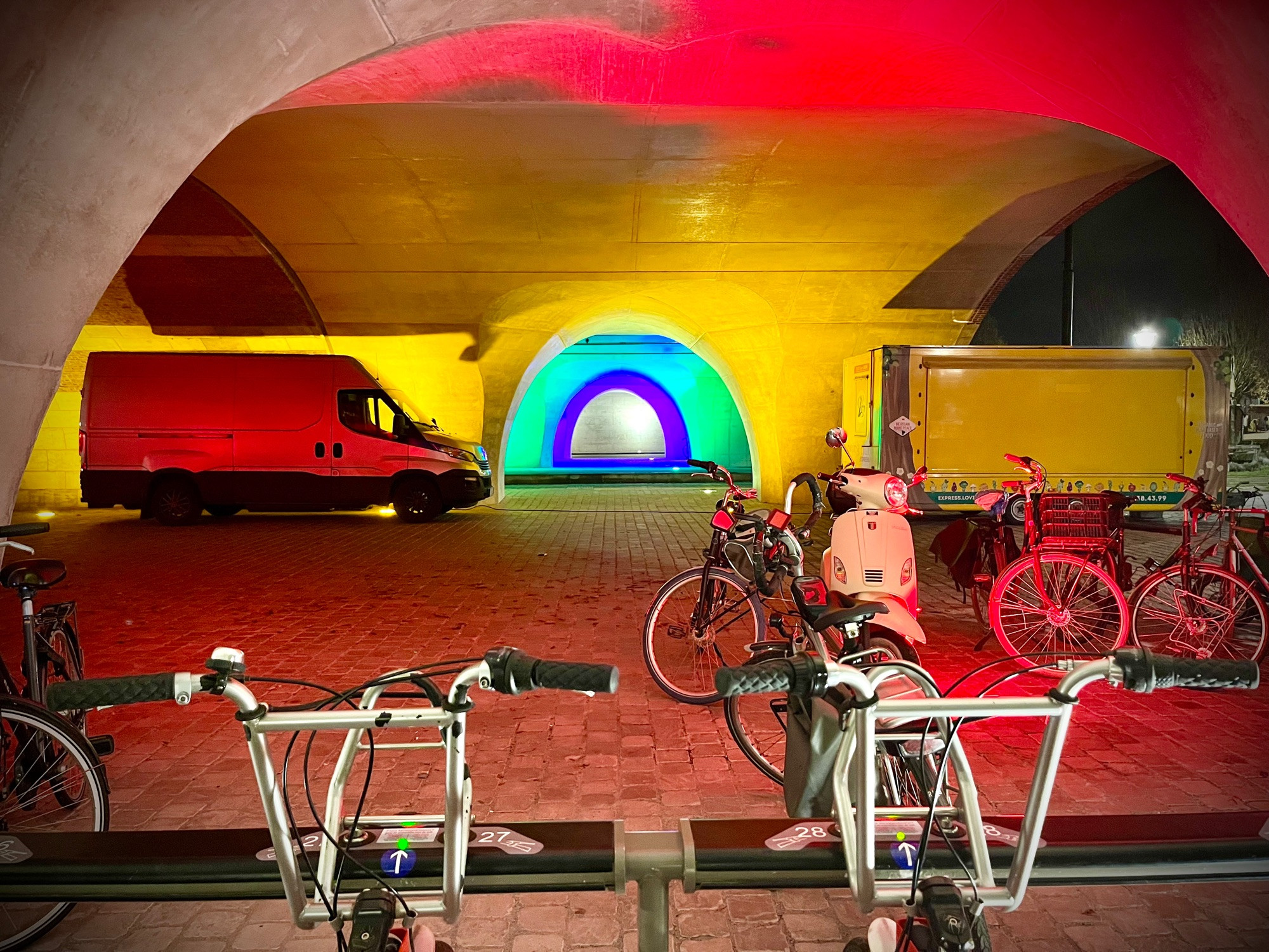 A colourfully lit spot beneath the railway bridge in the Antwerpen neighbourhood of Zurenborg. Bikes parked in the foreground; in the background is a red van. Lighting is mostly red, but there is a turquoise and purple rainbow shape on the far wall.