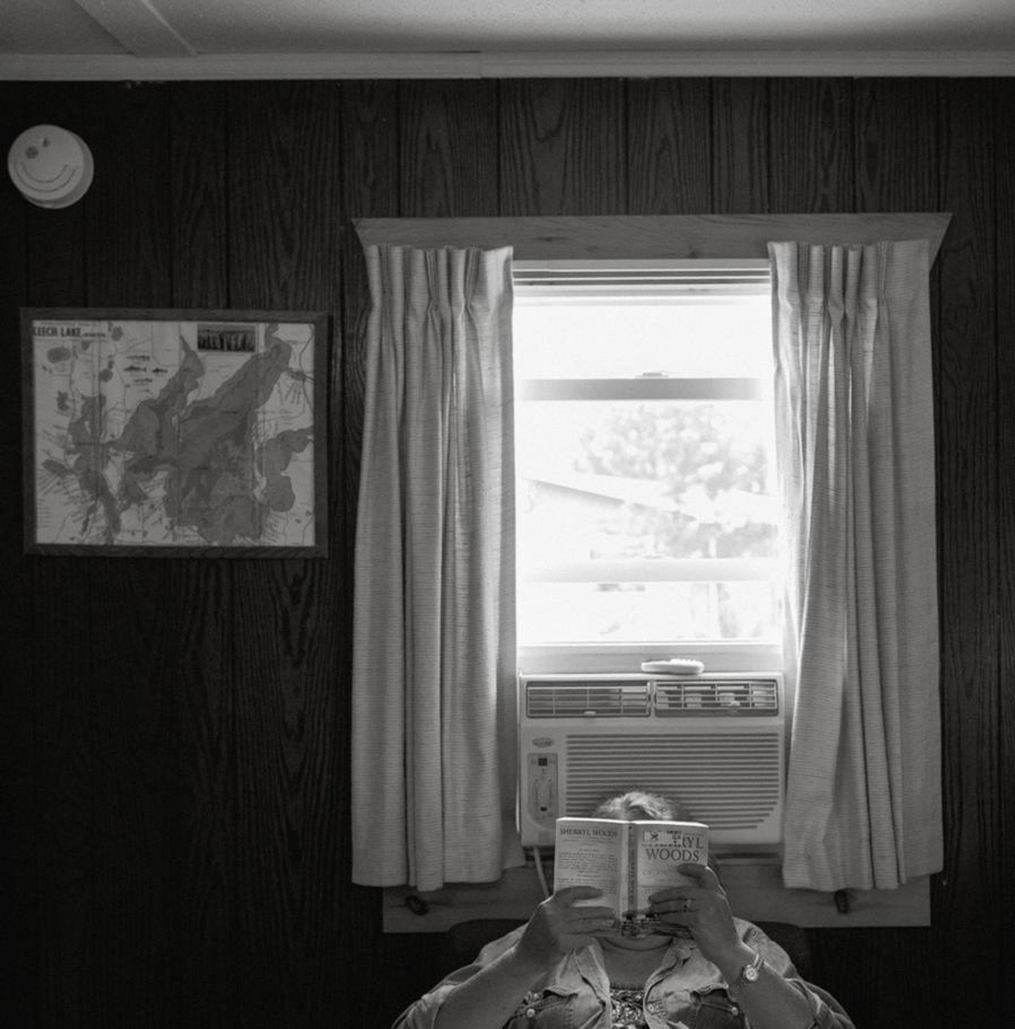 b&w photo of a woman sitting reading a book in a wood paneled room; she's underneath a window with the curtains open and an air conditioner behind her head. the book is obscuring her face. to the left of her is a smoke detector and a map on the wall.