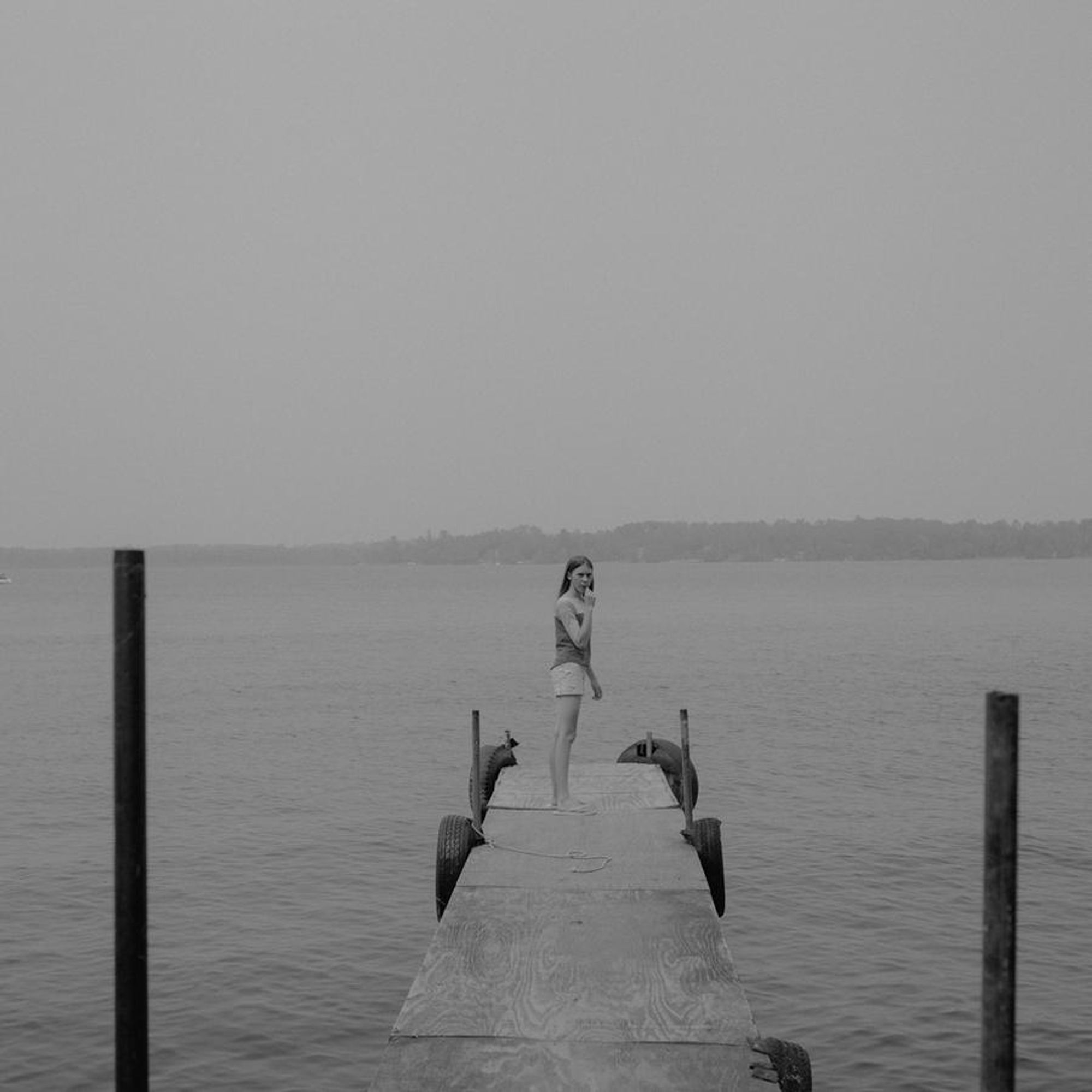 b&w photo of a young girl standing at the edge of a dock, biting her nails. the poles of the dock line up and almost line up with the horizon line created by the water/faraway land, which is centered in the frame.