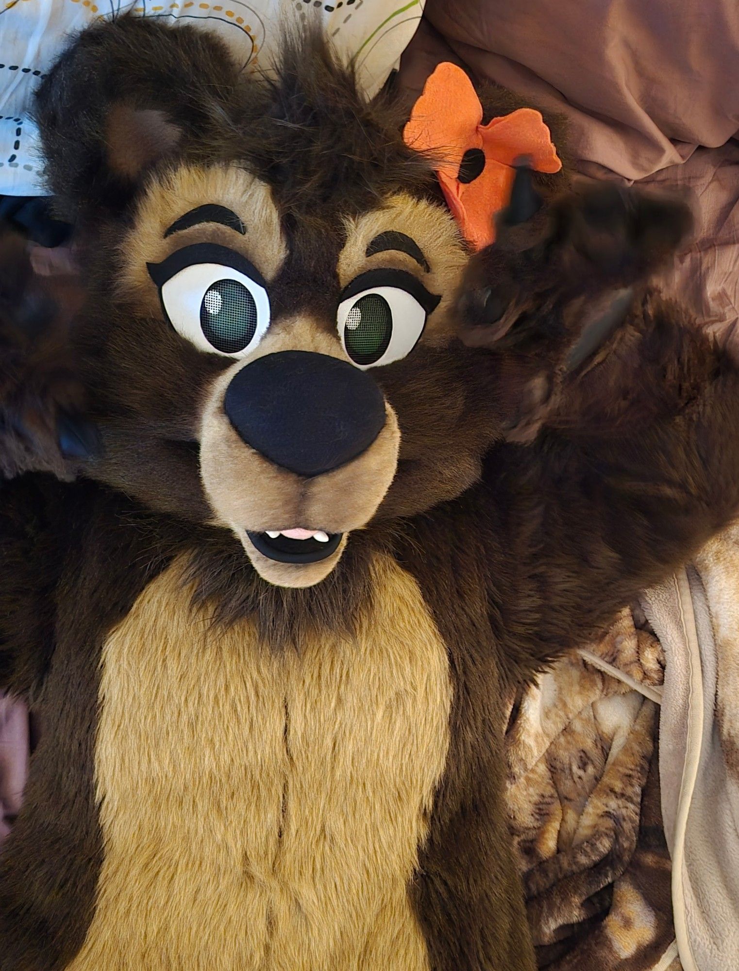 Poppy the bear fursuit lounging on a bed. She has a fabric Poppy flower attached to her ear.