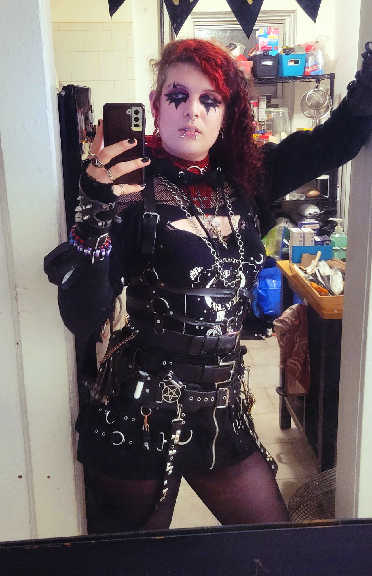 lilith in goth makeup and red hair, wearing a sweater cropped above the chest and a cami combining to make a boob window and a lot of belts.