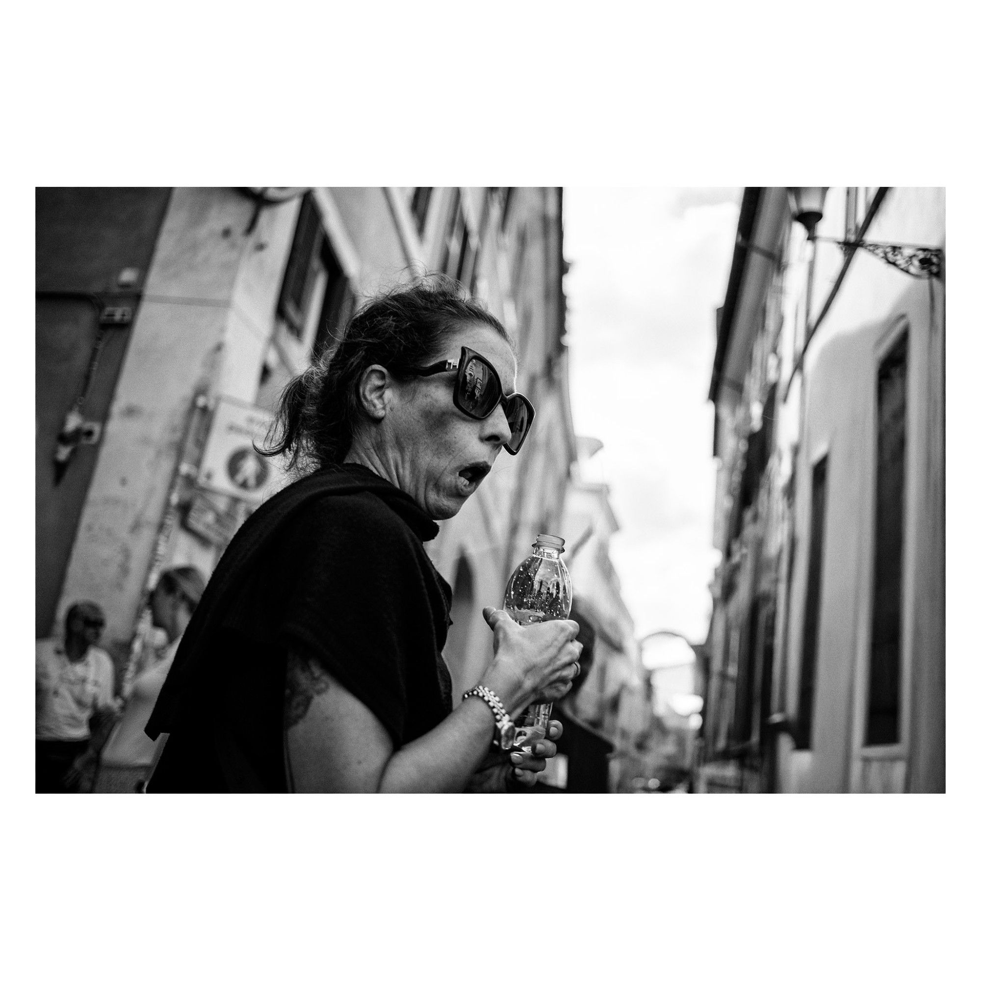 black and white photography of a surprised lady with black sunglasses holding a bottle of water in a narrow street. Roma