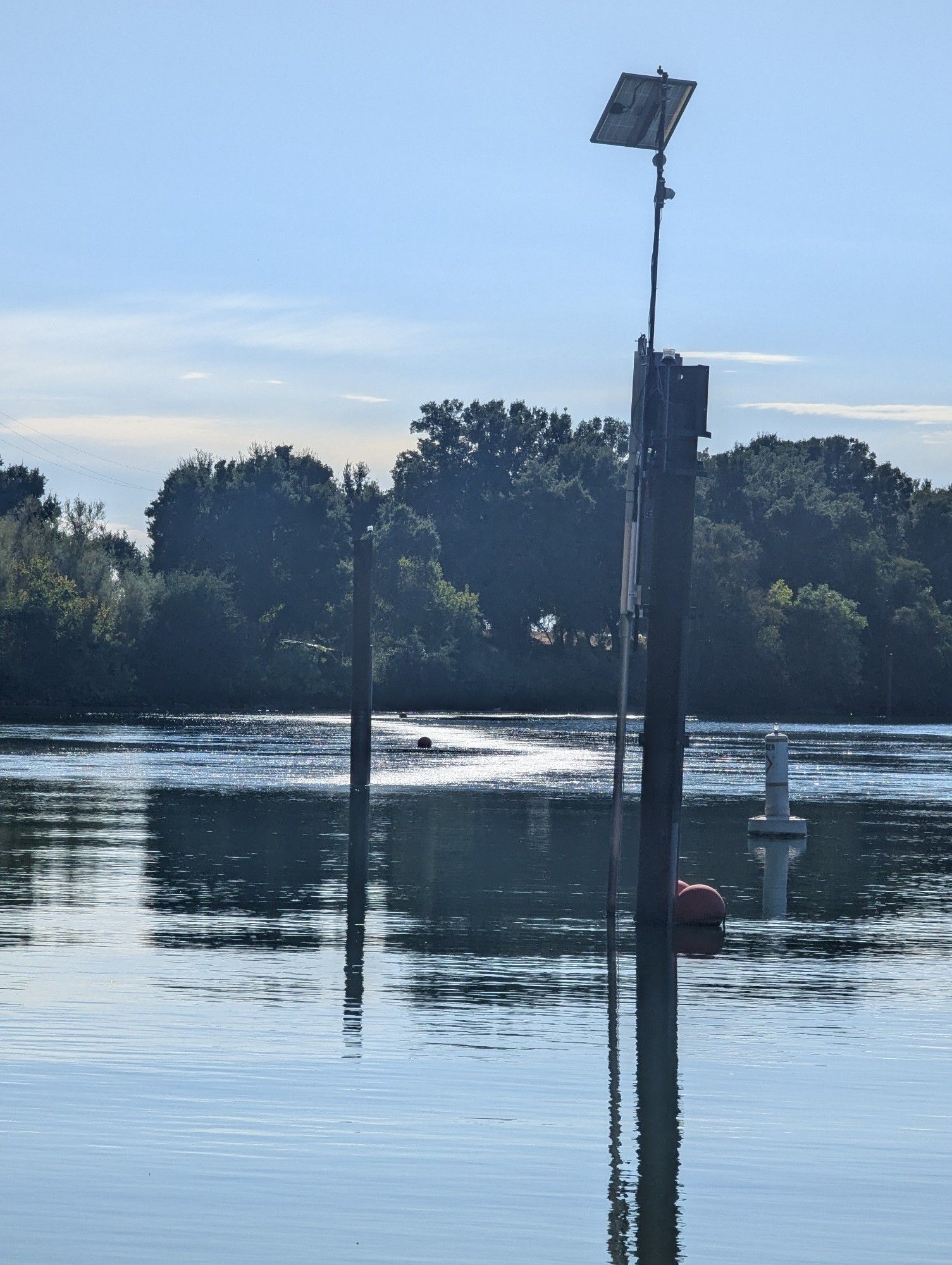 View of a snaking surface disruption along the top of a riverbed. Wooden towers rise from the river's surface, one with a solar panel used for blinking light at night. A heavily treed riverbank in the distance and a round red buoy and a white, upside down "T" buoy are in the middle distance. The river's surface is otherwise calm with some rippling; reflecting the nearly clear blue skies.