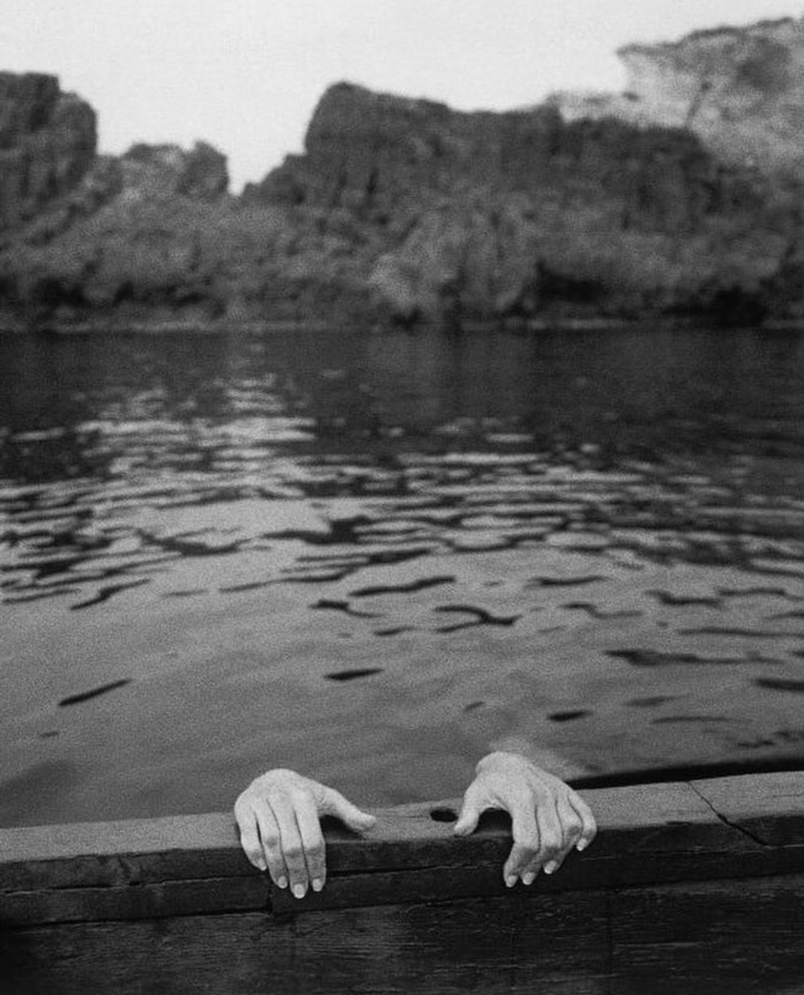 Black and white photo of two hands gripping the top of a low stone wall. The hands are reaching from a person who is in a body of water. The rest of the person isn't visible.