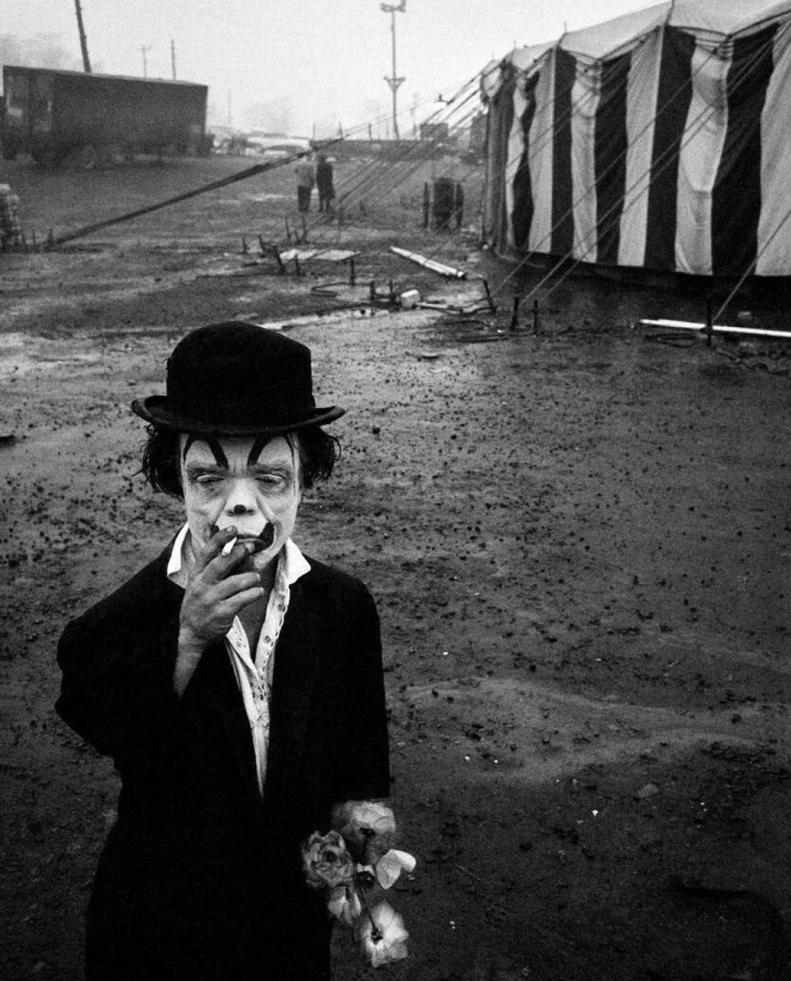 Black and white photograph taken by photographer Bruce Davidson. It features a little person in clown makeup smoking a cigarette. He is standing outside of what appears to be a circus tent.