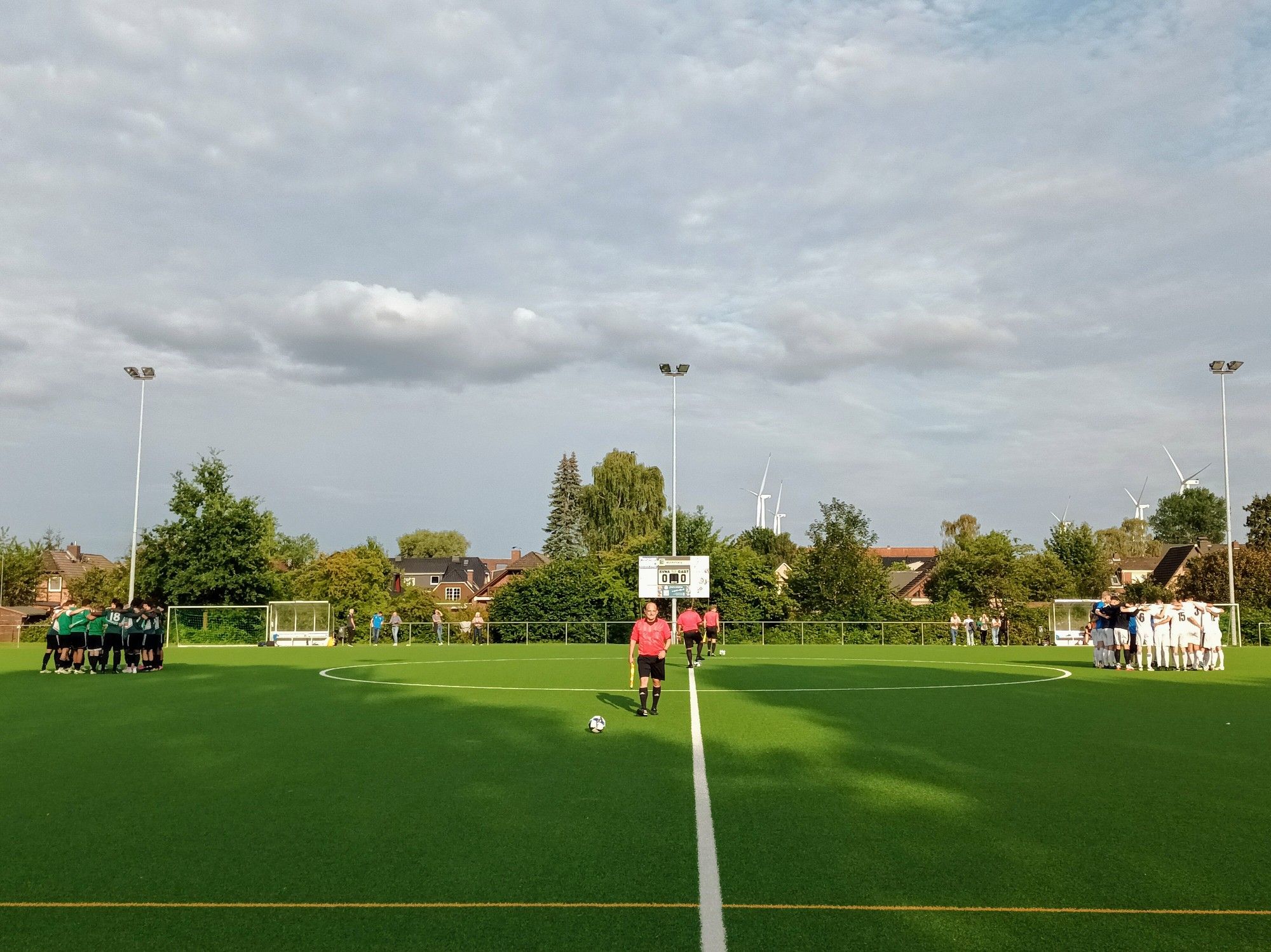 Mannschaftskreise und Schiedsrichtergespann kurz vor Anpfiff. Im Hintergrund 3 Flutlichtmasten und Windräder.