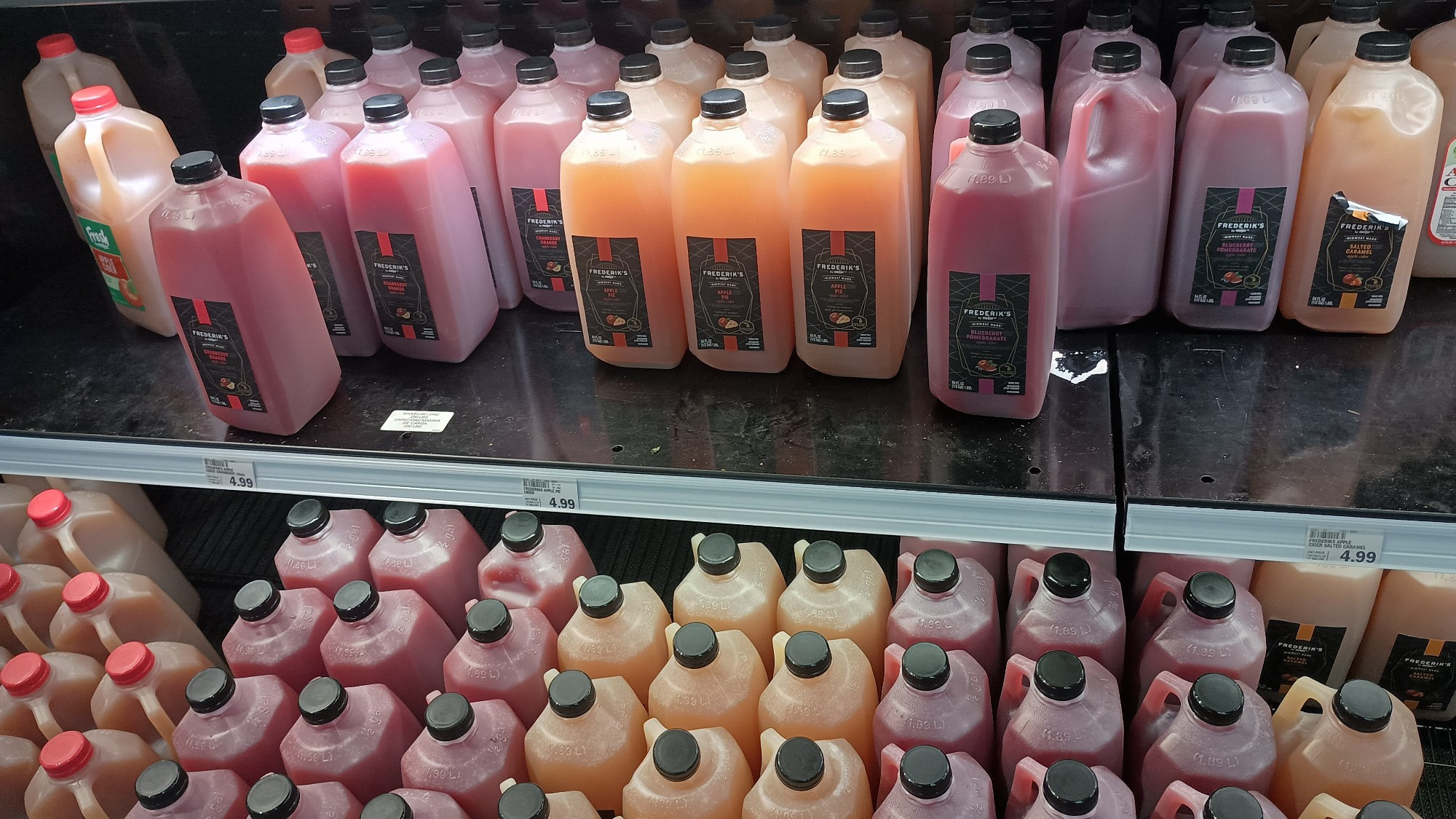 From left to right: apple cider, cranberry orange apple cider, apple pie apple cider, blueberry pomegranate apple cider, and salted caramel apple cider, arranged on a storefront cooler shelf.