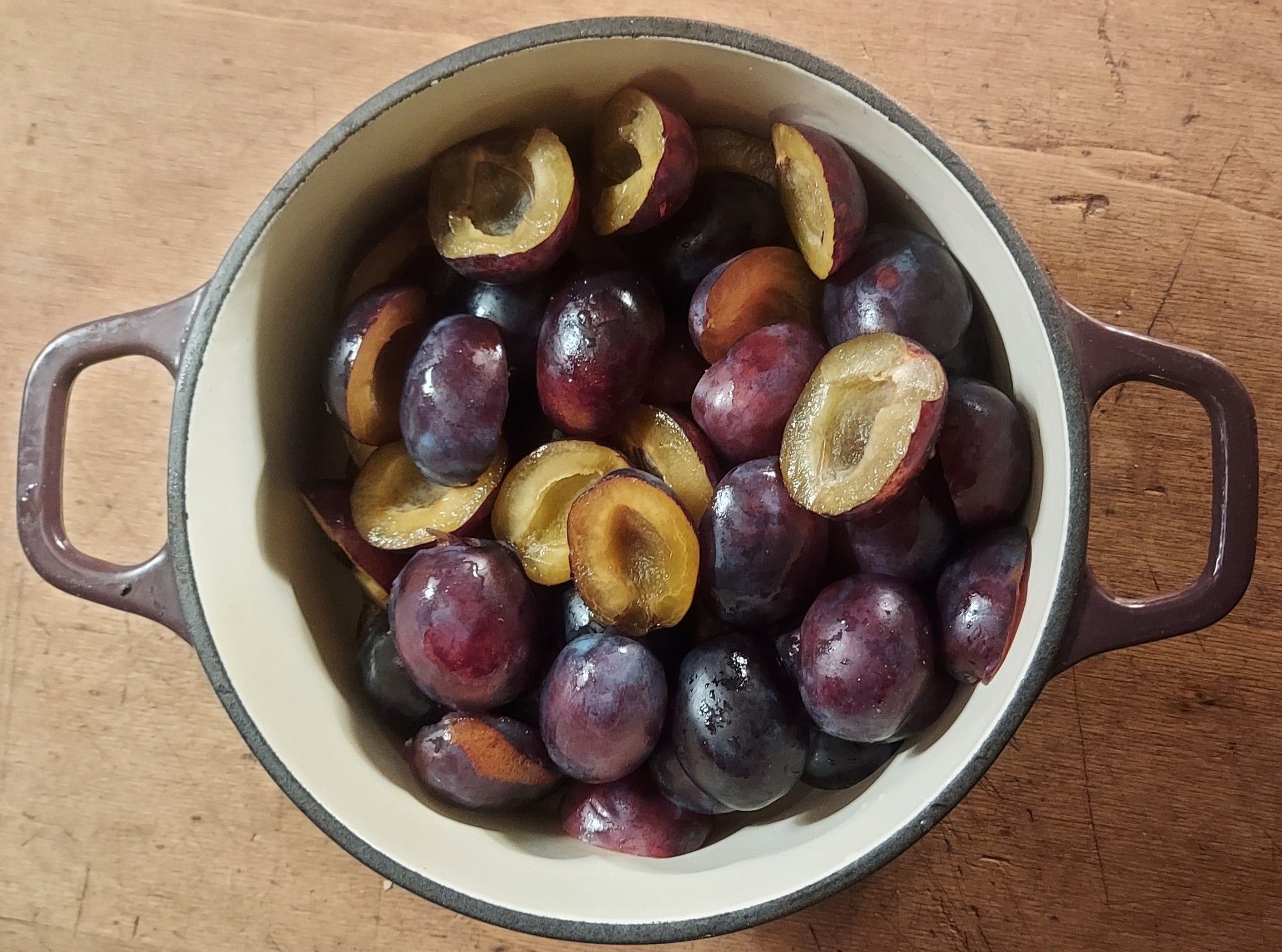 A round enamelled cast iron cooking pot containing halved purple-skinned plums showing their golden yellow flesh.