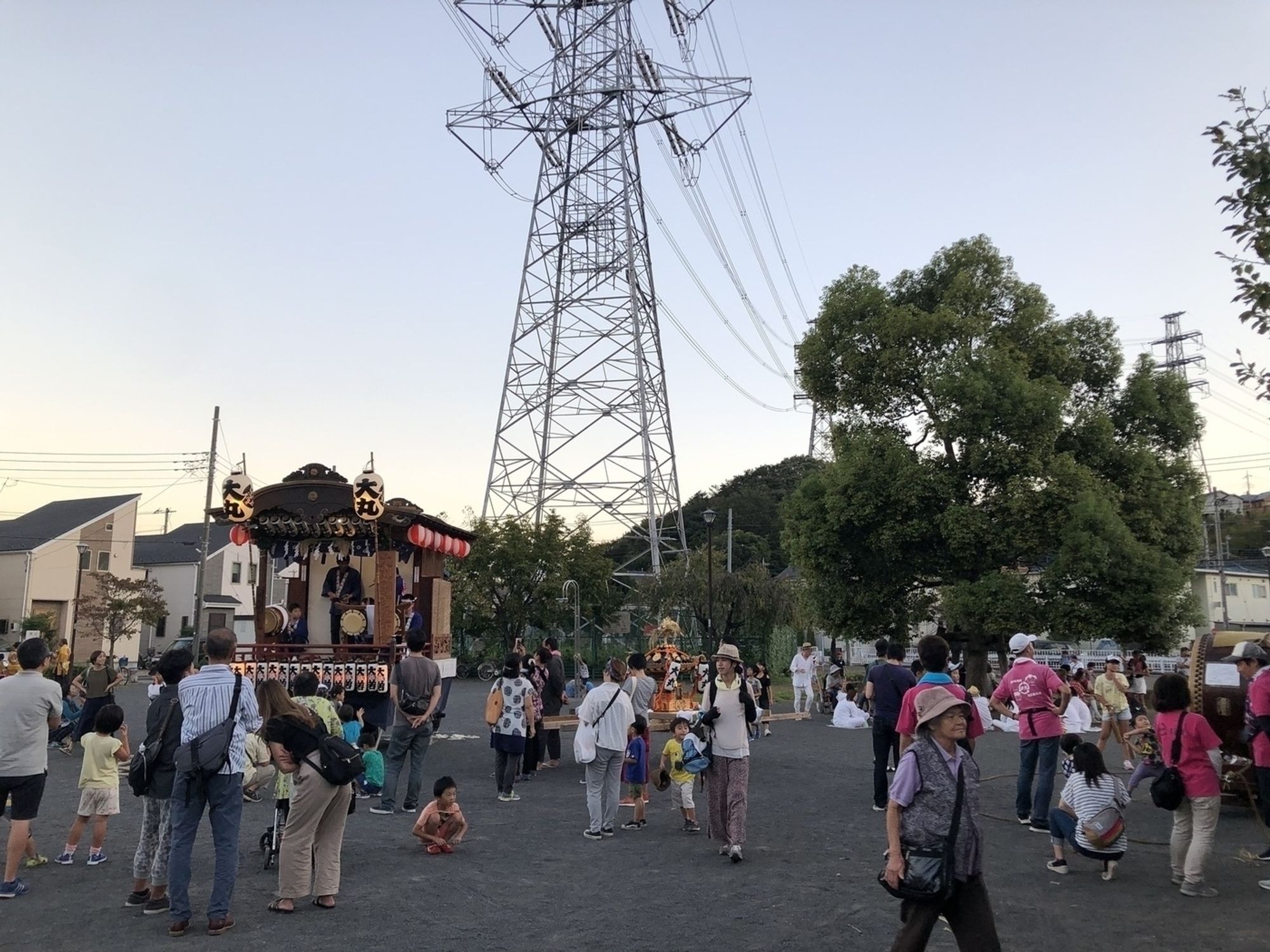 People in a Japanese neighborhood are gathered in an open area near a festival float and a large power transmission tower.