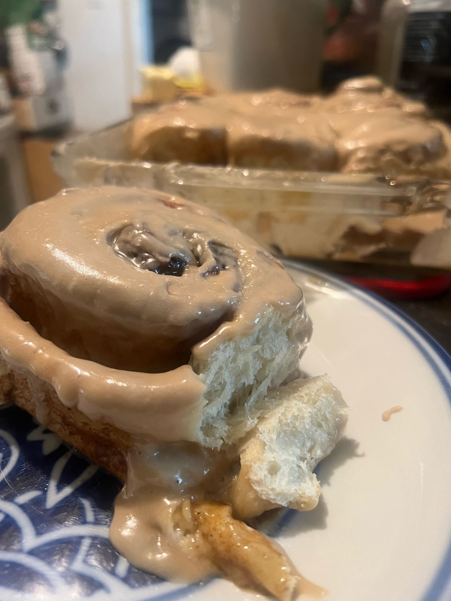 Photo of a single cinnamon roll on a plate with the rest of the tray in the background. A piece of the roll is broken, showing a light fluffy interior. Light brown maple glaze drips down the sides of the roll.