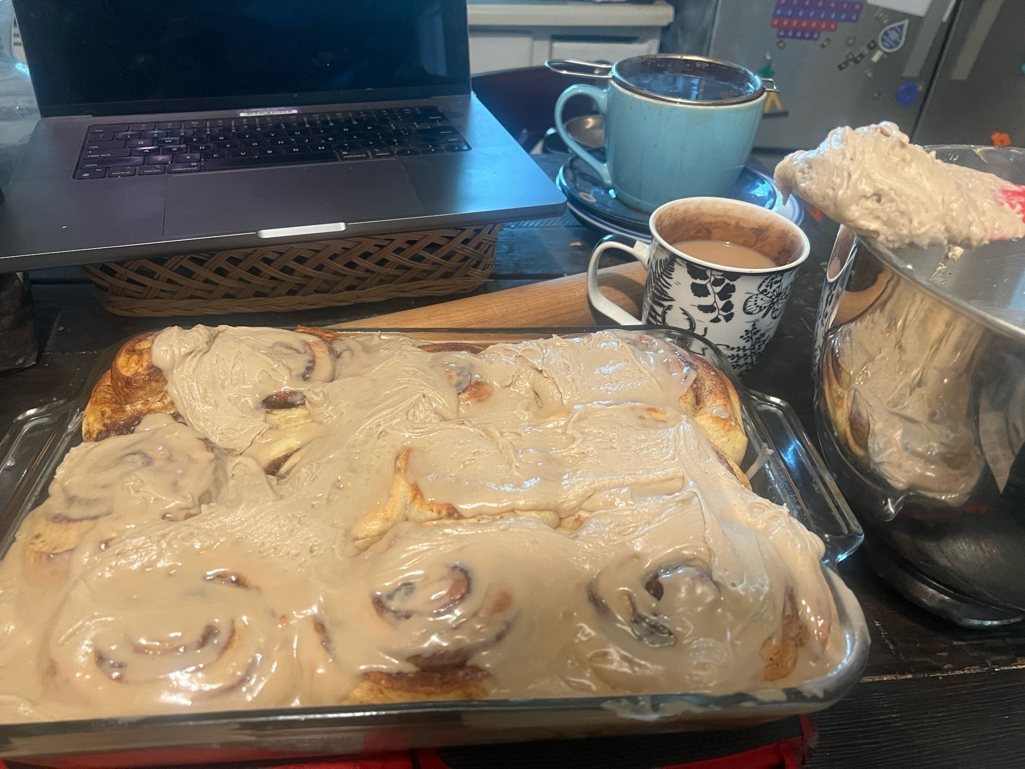 Photo of a full tray of cinnamon rolls, covered with a light brown maple icing. A mug of tea and a computer sit behind it, and a mixing bowl with icing spatula is to the side.