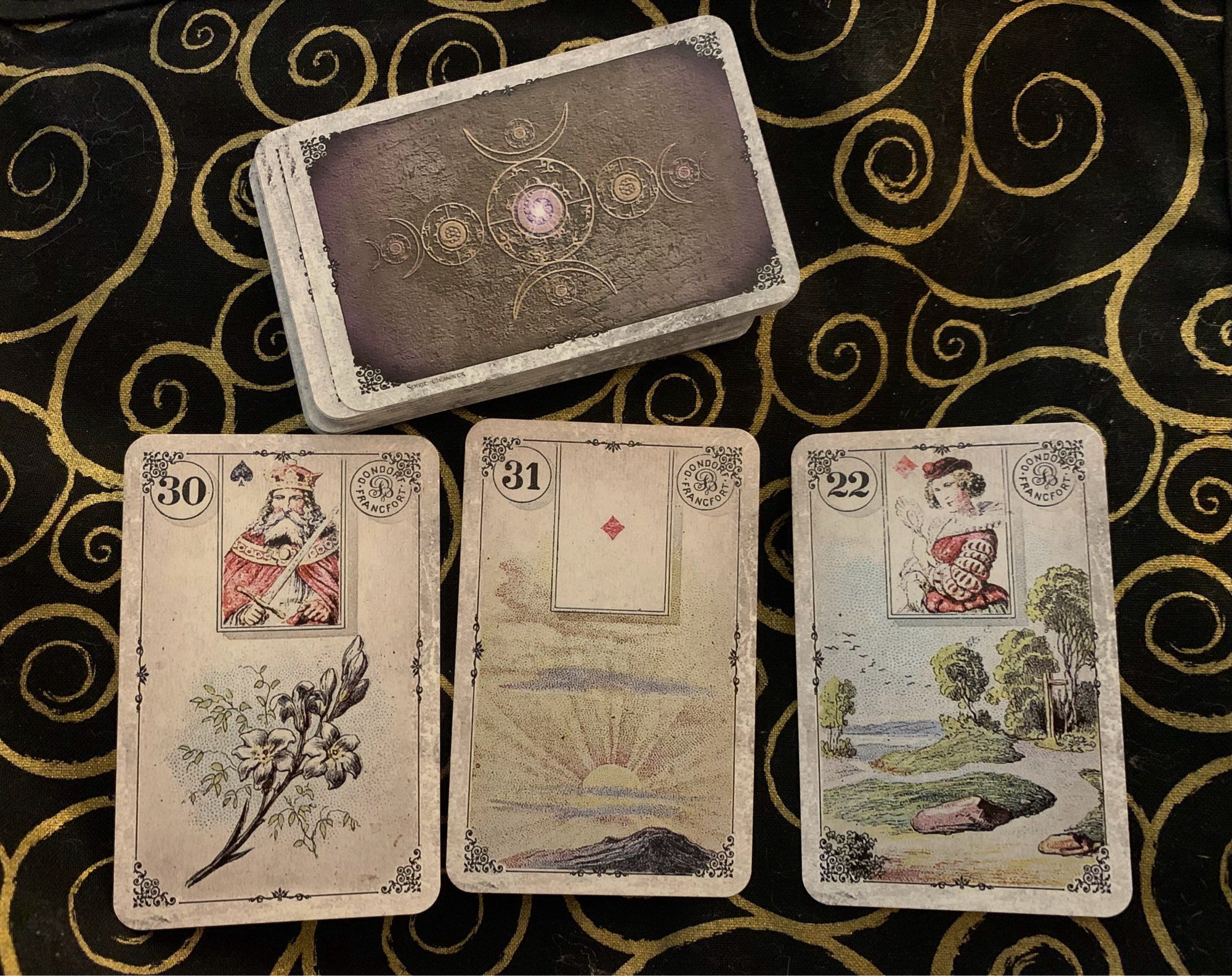 Photo of a three-card Lenormand pull from the Dondorf 1910 Reproduction pack on a black altar cloth with gold spirals, showing from left to right the Lilies, the Sun, and the Crossroad. The remainder of the cards are placed above these three.
Apologies for the cards not being straight, but Drayko (the grey tabby cat) decided to jump on it as I took the shot.