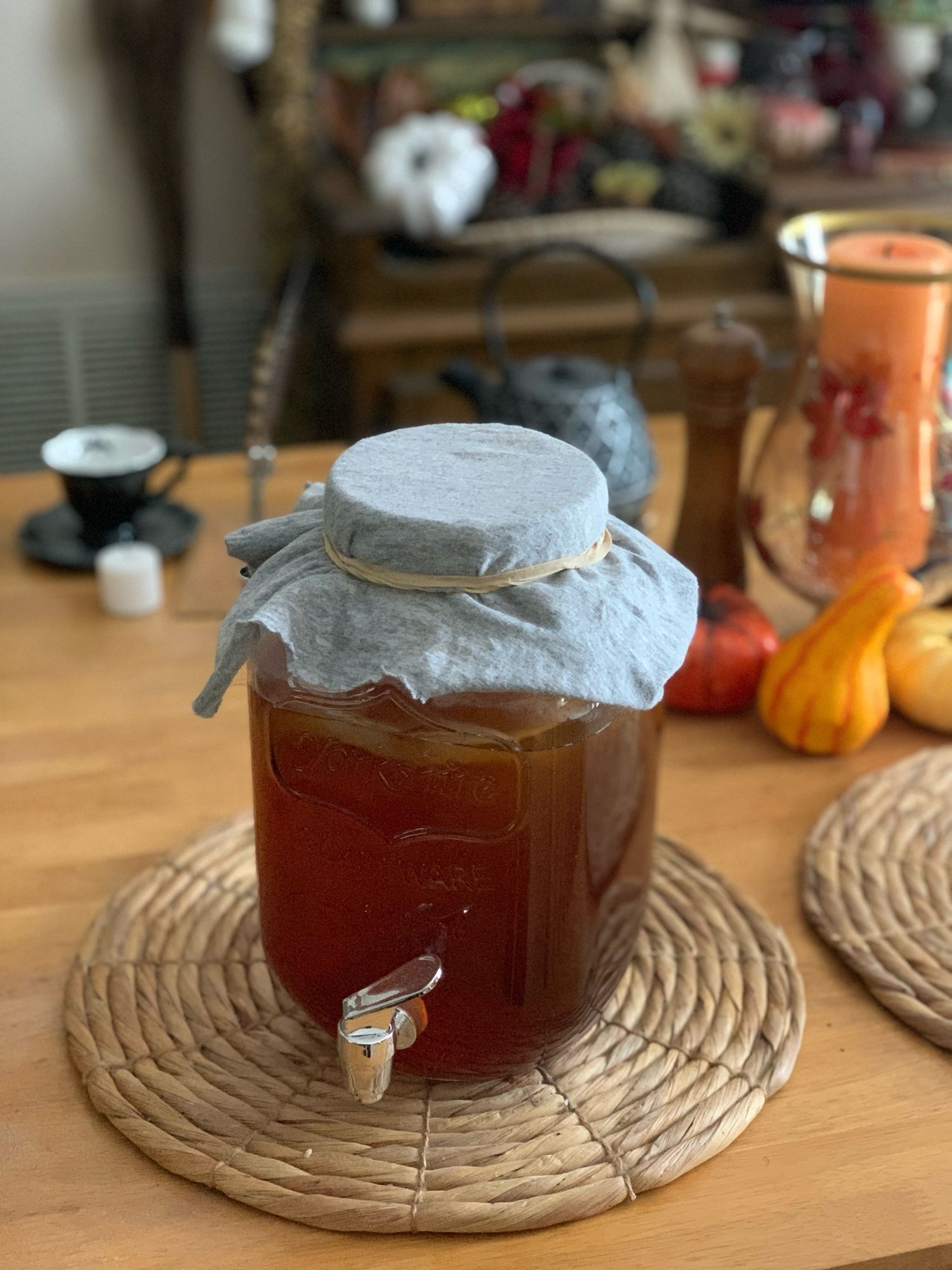 Photo of a gallon jug of freshly brewed Kombucha ready to set aside for several weeks for fermentation. 

Blurred in the background is the wooden table it’s resting on, a black with bats bone china teacup and saucer, a white votive candle, a hawk quill pen, a cast iron teapot, a glass hurricane candle holder with a large orange pillar candle, a few small pumpkins and gourds.