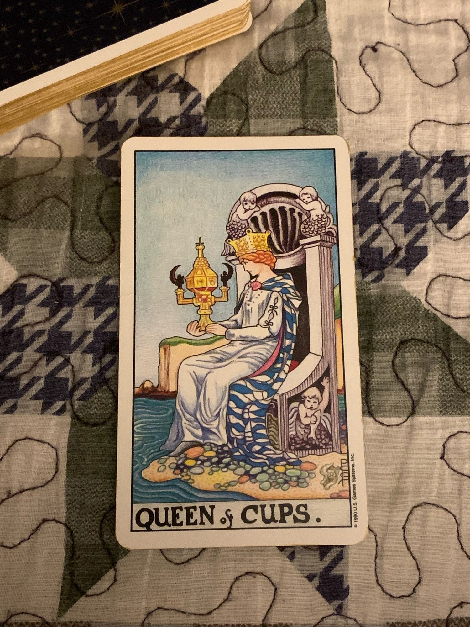 Photo of Queen of Cups from Universal Waite Tarot. 
The Queen of Cups is a beautiful, introspective woman who sits on a throne at the edge of the sea. In her hands, she holds a golden, cup adorned with handles shaped like angels. Unlike most of the cups in the Suit of Cups cards, this cup is closed – showing that the Queen’s thoughts and feelings come from her subconscious mind and the depths of her soul.
She sits upon a stone throne decorated with images of sea-nymphs, fish, and scallop shells. The sea and fish are symbols of the unconscious mind, and water represents emotion, spirit and perception. A cloudless, bright-blue sky and placid water of the sea surround her. Her feet do not touch the water; they rest on colourful pebbles on the shore. It is as if she is connected to her emotions (as represented by the water), but not overwhelmed by them.