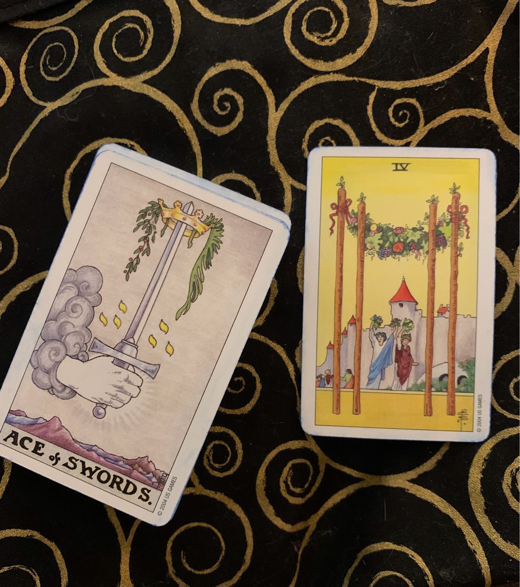 Photo of the Four of Wands from the Mini Universal Wait Tarot to the right, and the Ace of Swords as the “Shadow” card on the bottom of the deck, to the left on a black altar cloth with gold spirals.