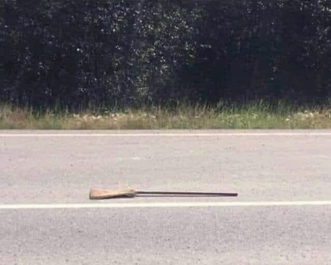 Photo of a road with a broom laying in the far lane. Trees are in the background on the other side of the road. 