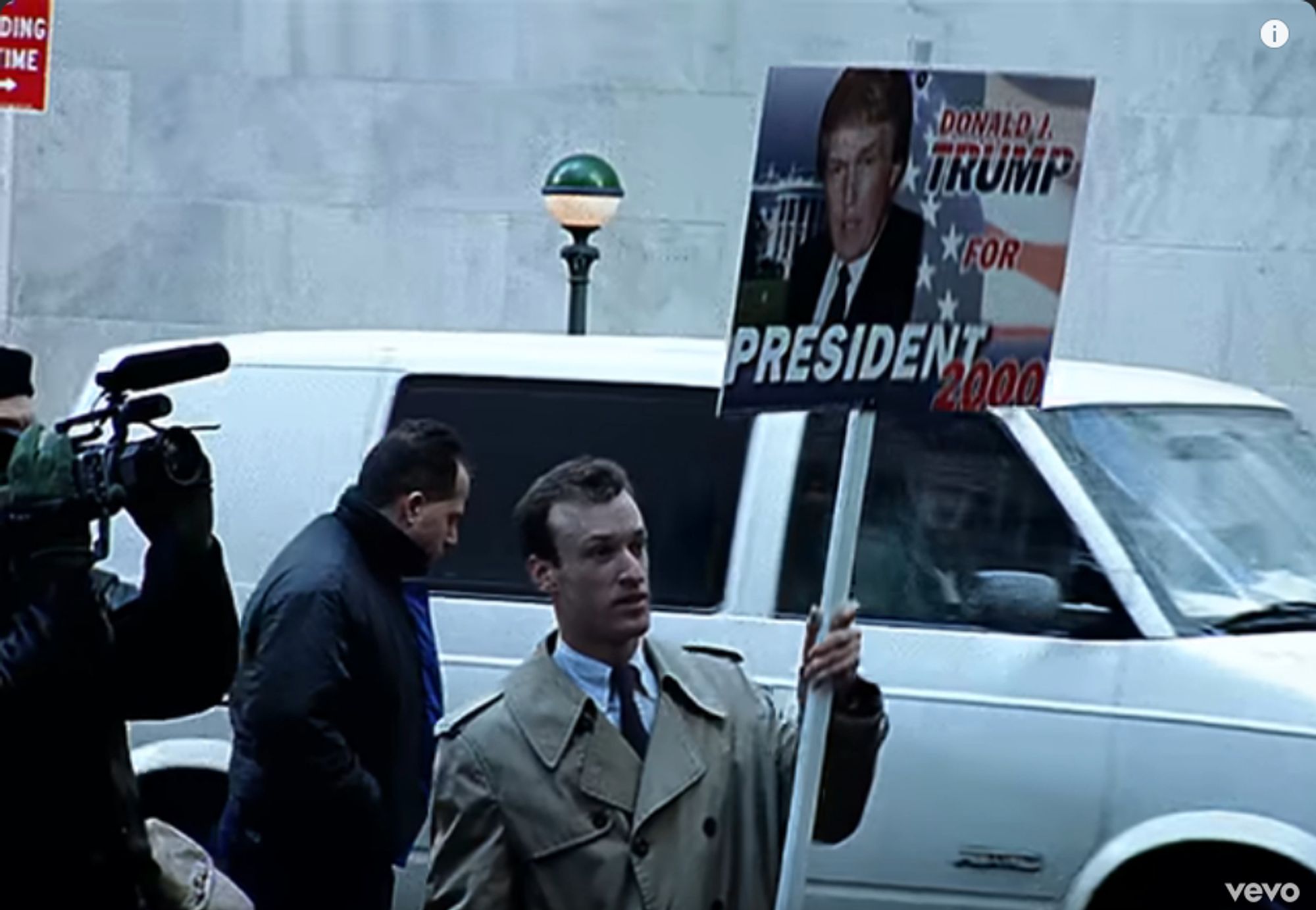 a guy holding a sign saying "donald j trump for president 2000" with an image of donald j trump in front of the white house and the american flag. behind him there's a car and a guy walking, and someone filming him, it doesn't look staged at all