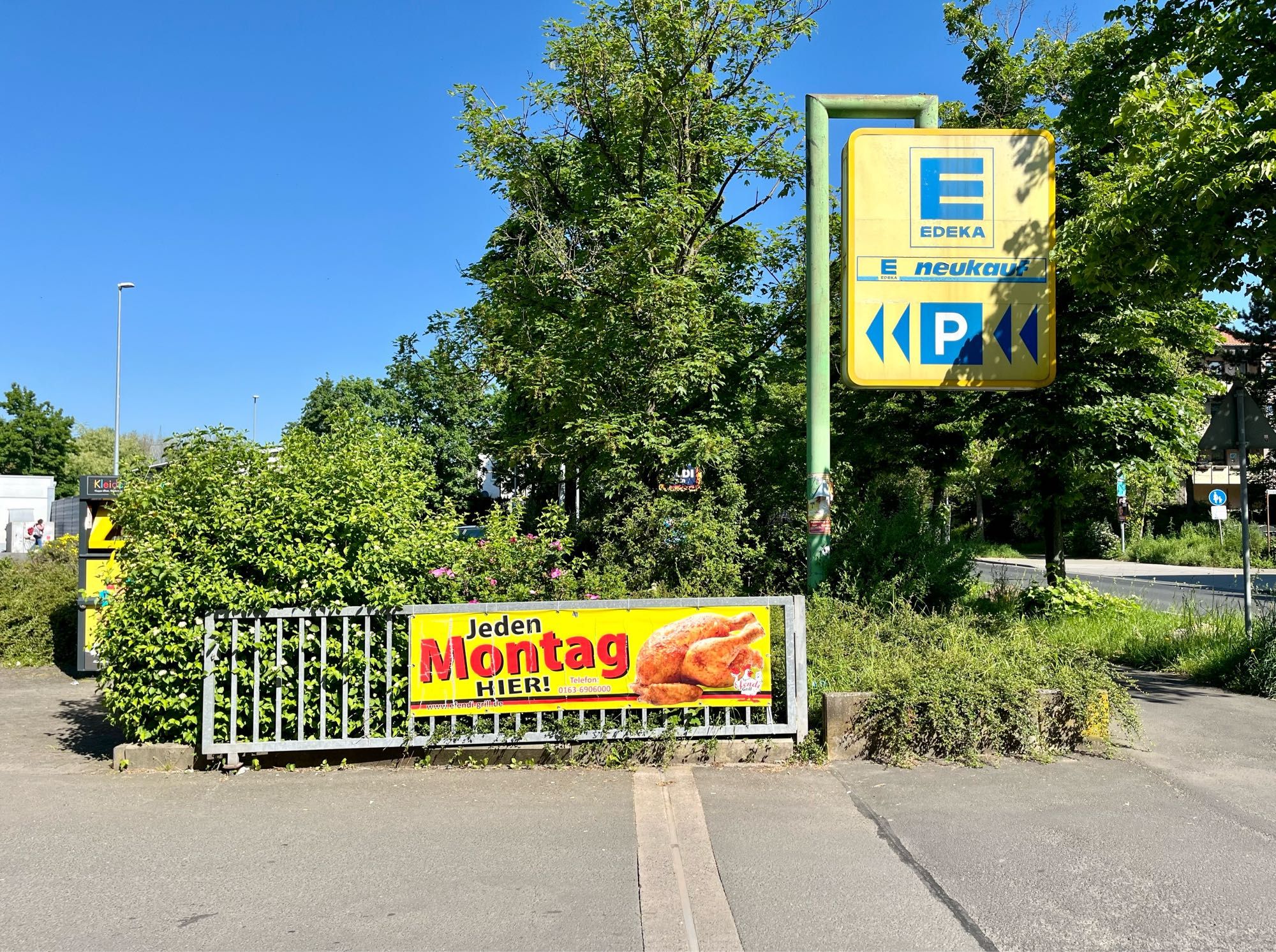 Am geöffneten Einfahrttor zum Supermarkt-Parkplatz hängt eine gelbe Reklame mit roter Beschriftung, auf den ein Grillhähnchen zu sehen ist, und die darauf hinweist, dass es jeden Montag diese Hähnchen gibt. Rechts davon sieht man ein gelben Hinweistafel mit blauer Schrift, die auf den Edeka Parkplatz hinweist. Alles ist Eingebettet in viele grüne Bäume und Sträucher. im Vordergrund sieht man grauen Asphalt. Über allen ein strahlend blauer Himmel.