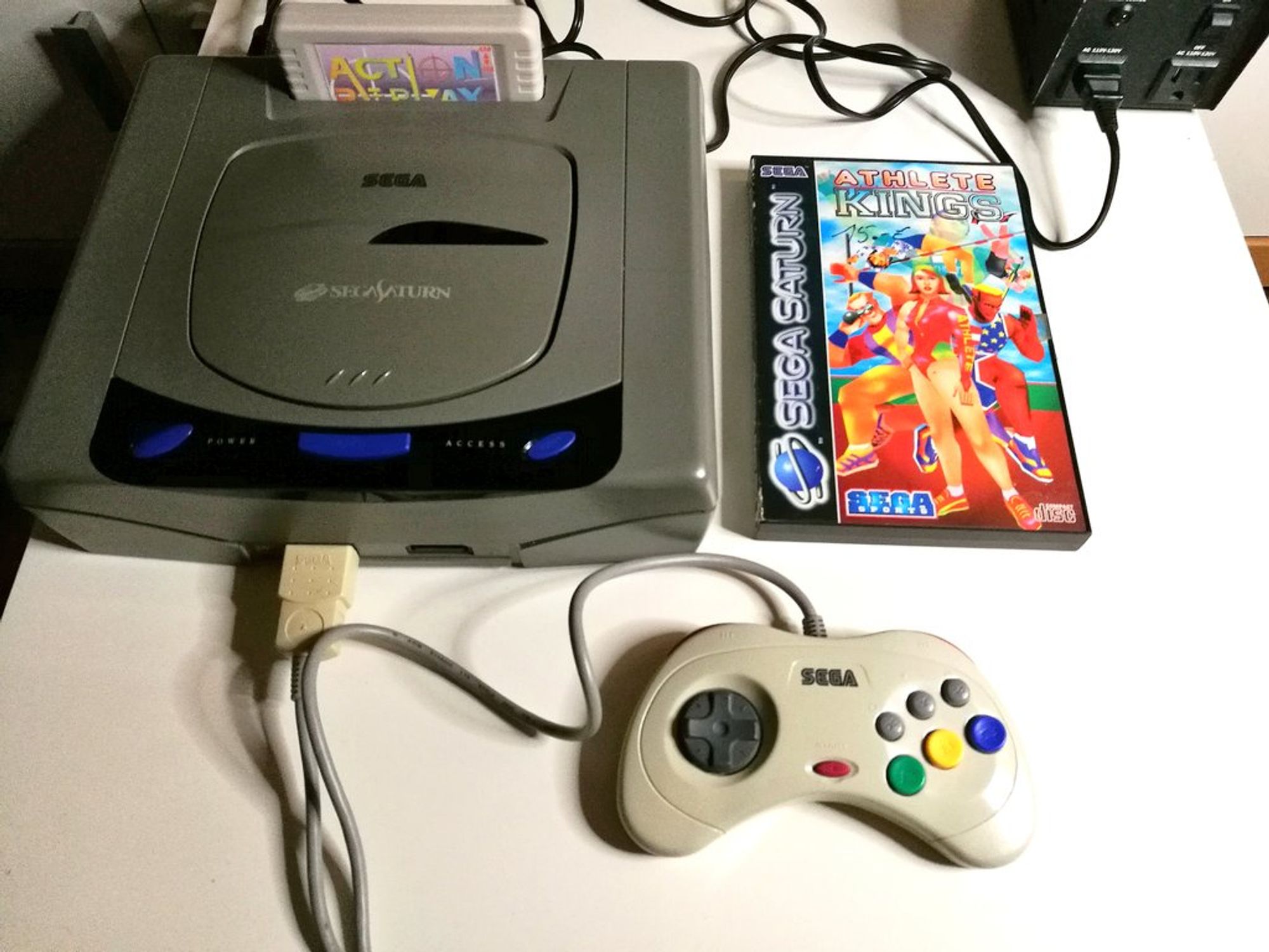 Links ein grauer japanischer Sega Saturn mit weißem Controller und Action Replay Modul. Rechts Athlete Kings als Pal Version für den Sega Saturn.