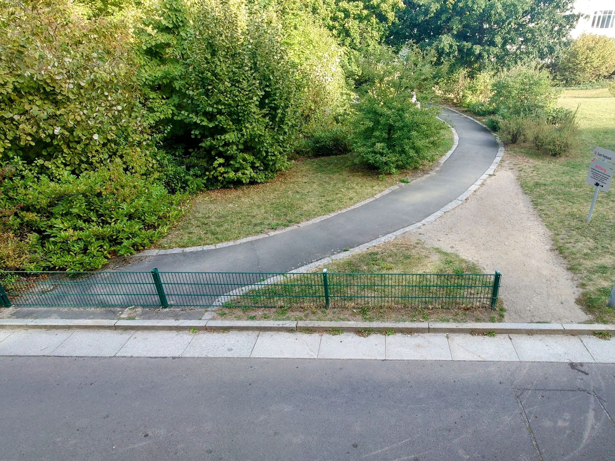 Ein asphaltierter Weg wird durch einen flachen Zaun begrenzt. Daneben ist ein Trampelpfad.