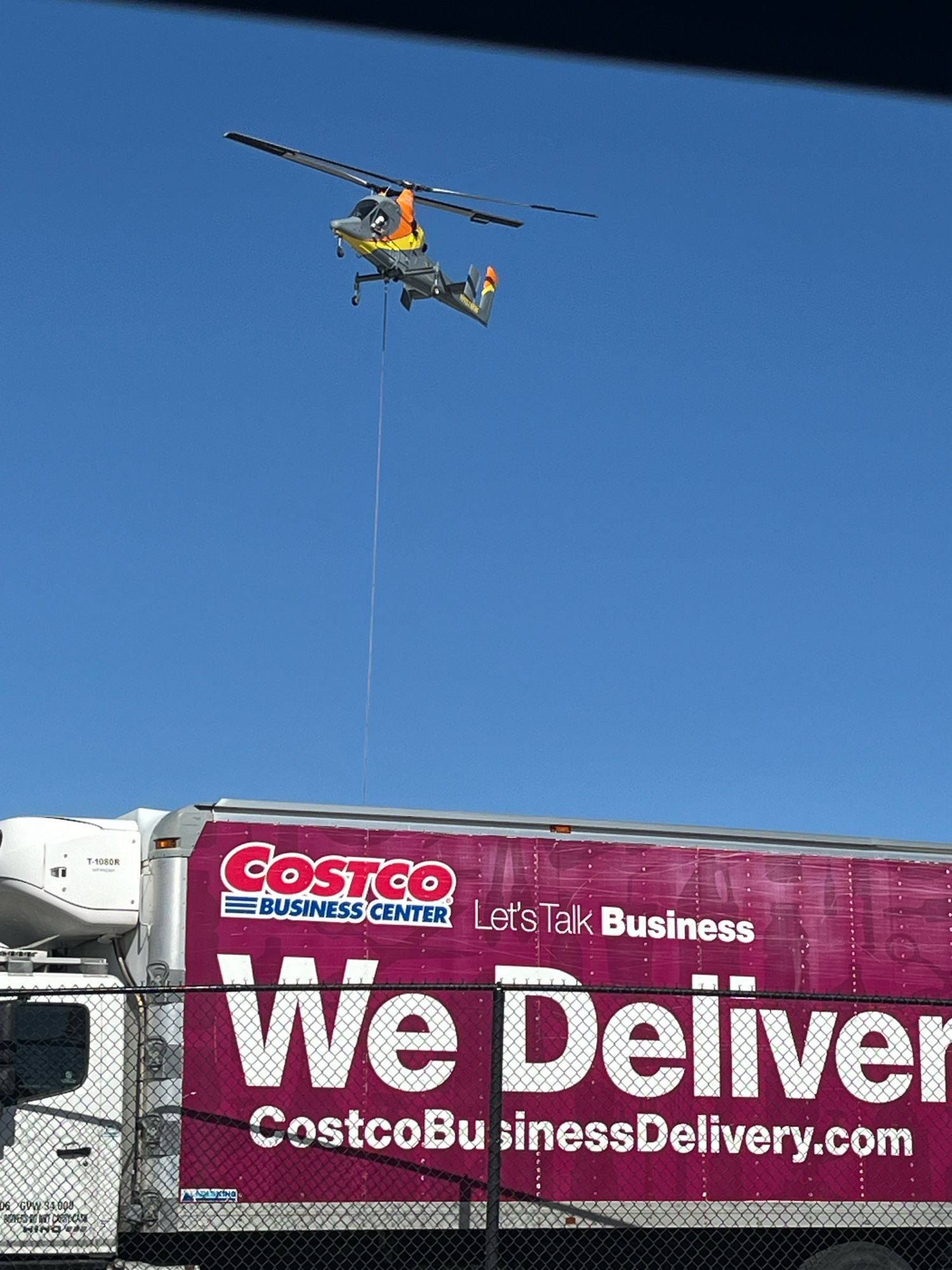 Helicopter hovering low with descended line over Costco truck with “we deliver” logo on the side.