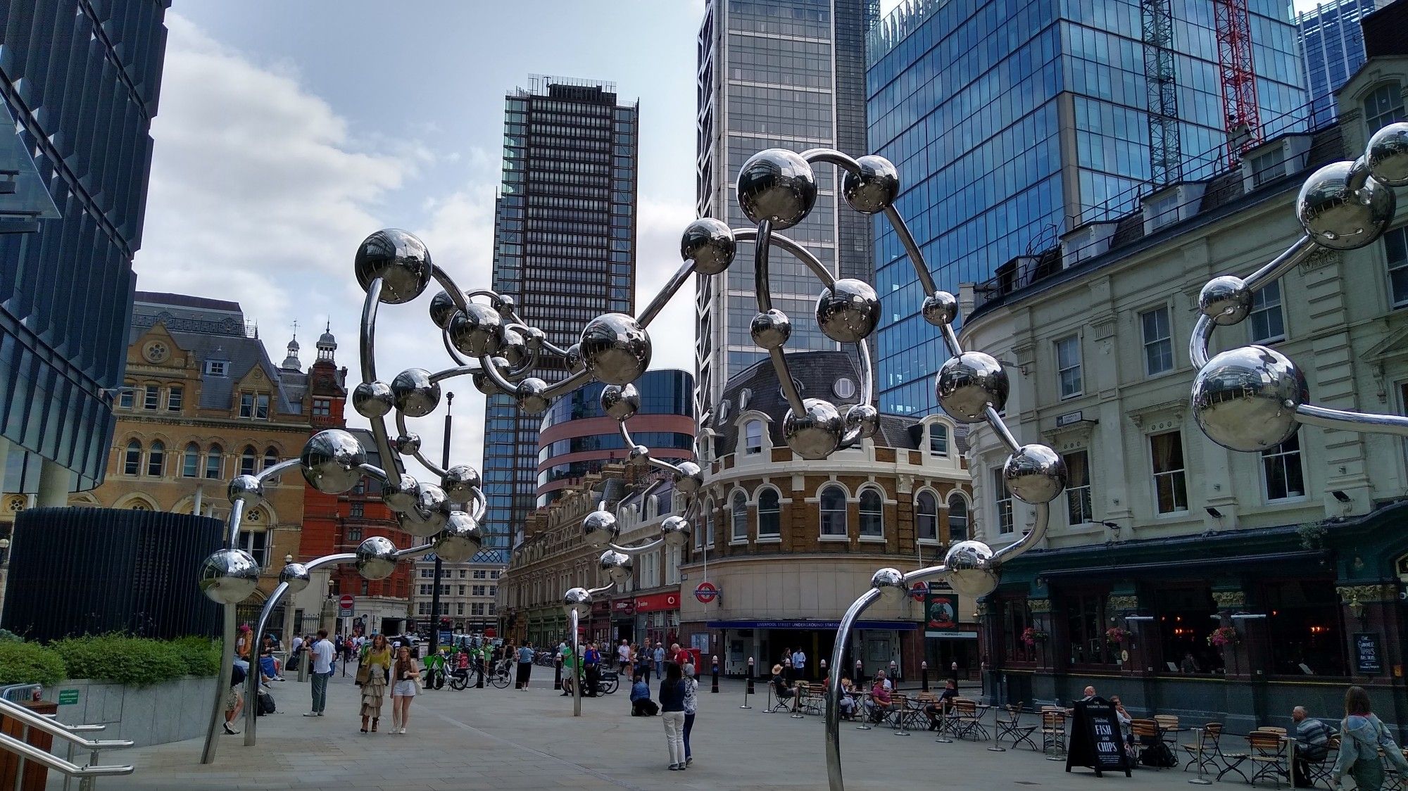 A towering structure of reflective balls held together with what looks like reflective strings, but on a grand scale. They start near the ground and rise up and down in waves. There are skyscrapers in the background.