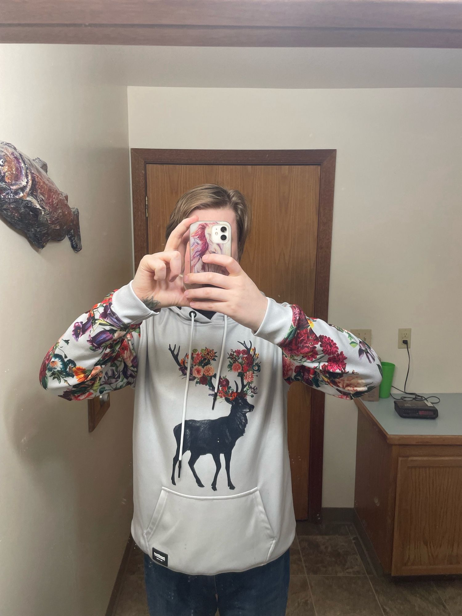 A mirror selfie of a white hoodie with a black silhouette of a stag. The stag’s antlers are blooming colorful flowers and the sleeves are covered in the same flowers