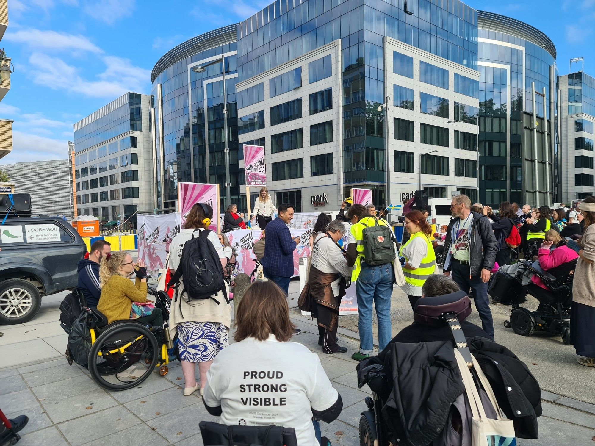 Un groupe de personnes debout et en fauteuil roulant devant une estrade de la Freedom Drive, bâtiments du quartier européen de Bruxelles dans le fond