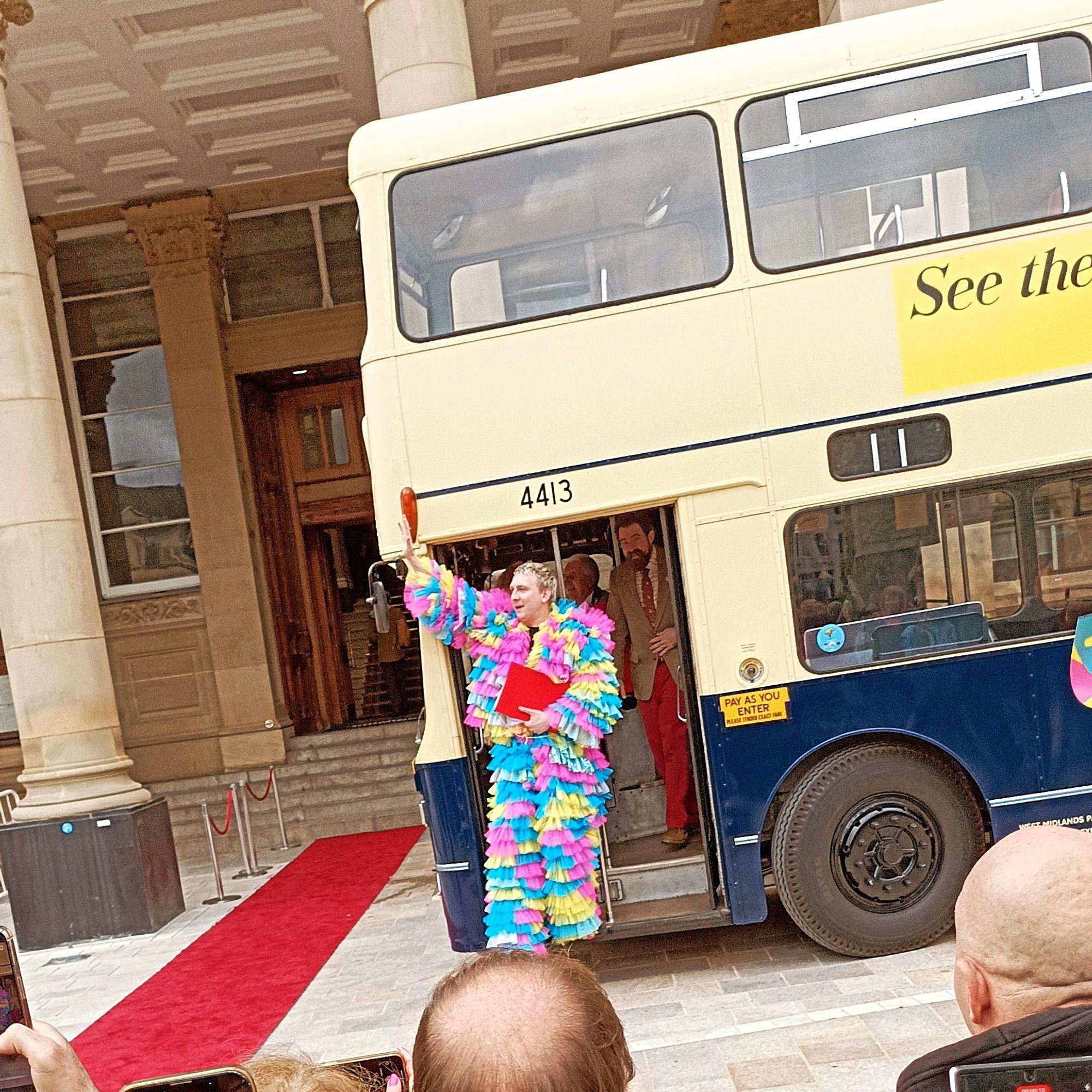 Joe Lycett waving from the platform of a number 11 bus outside Birmingham Council House