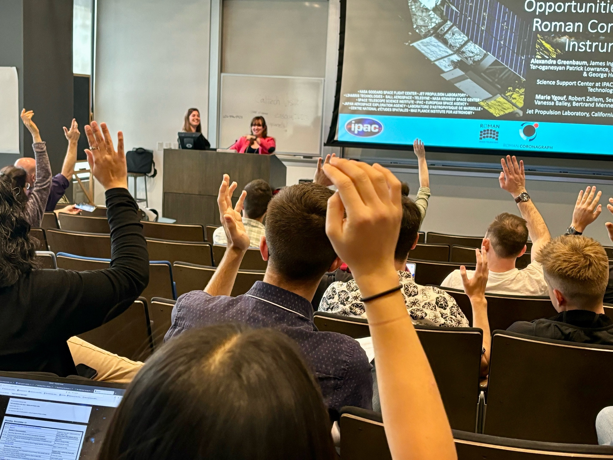 Emily and Catherine behind a podium facing an audience with their hands up.