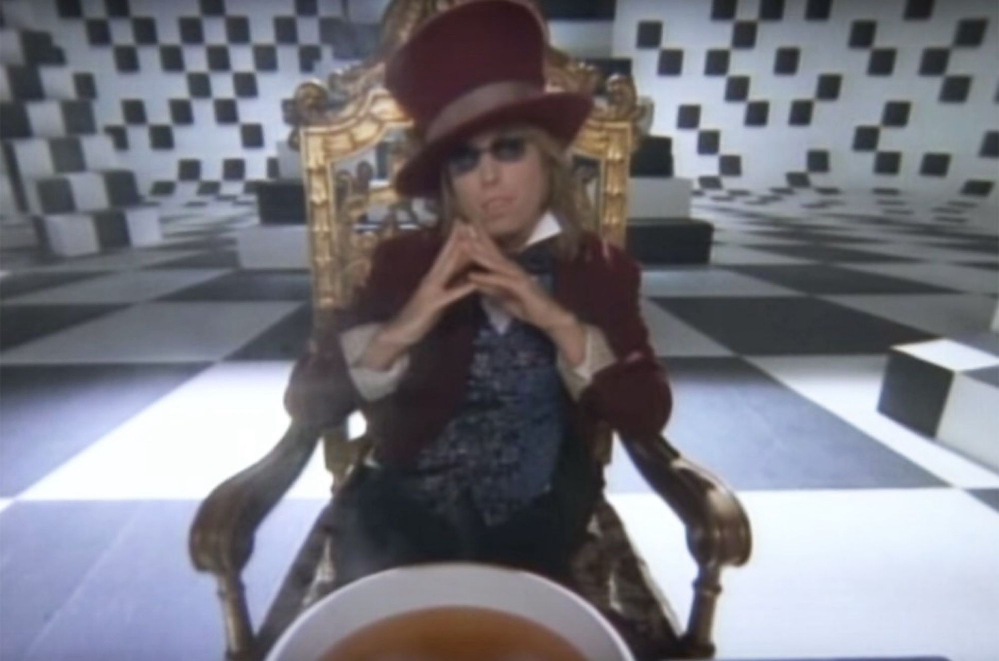 Image of a large room with black and white checkerboard floor and matching checked walls. In the foreground, Tom Petty is sitting in an ornate chair and dressed as the Mad Hatter character from Alice in Wonderland.