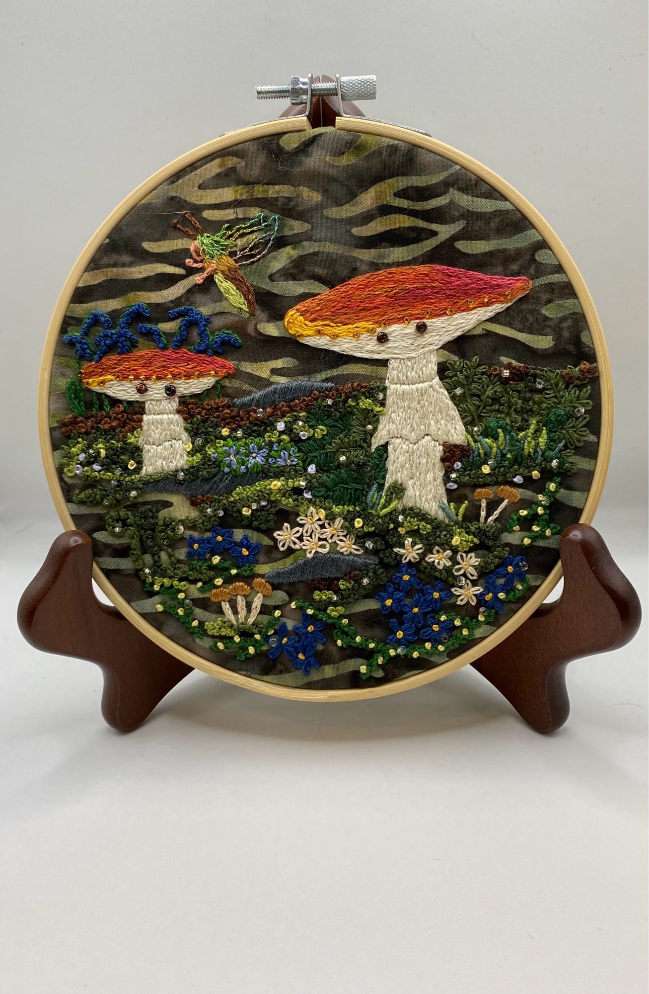 Multimedia embroidery with beads. Two amanita Muscaria mushrooms with little bead eyes on green camouflage cotton material surrounded by smaller mushrooms, ferns, and blue flowers. Framed by a wooden hoop and perched on a wooden plate stand.