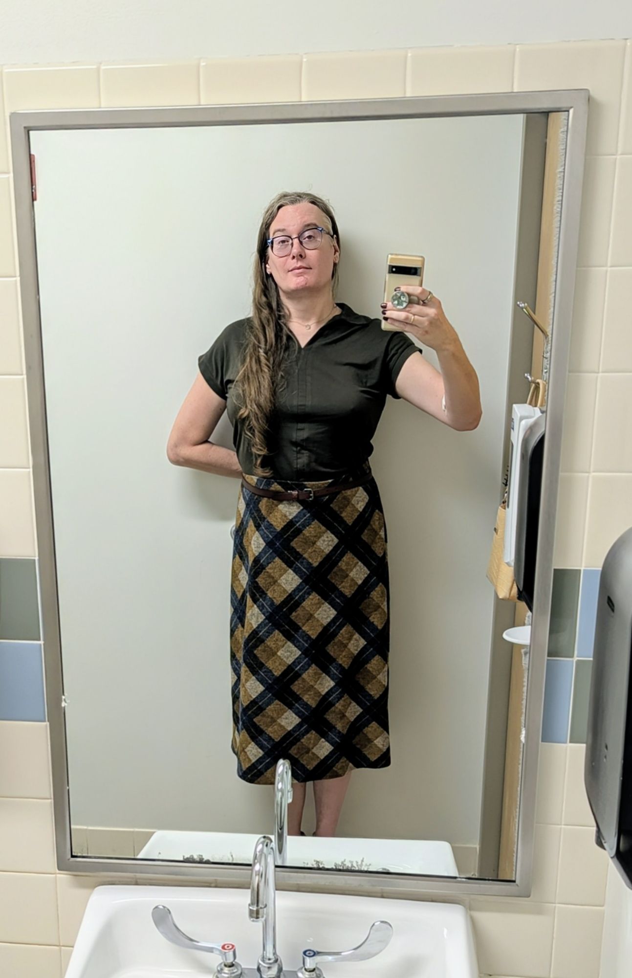 Saga taking a mirror selfie. She's wearing a dark green collared short sleeve shirt and a flowy plaid dress, beige/tan/navy/dark green, that hits at mid-calf.