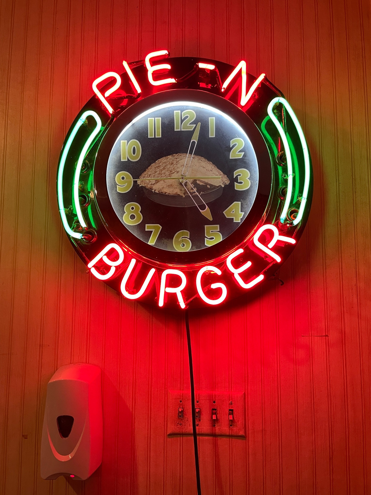 Round neon sign with analog clock, center of clock is a pie. Surrounding clock are letters spelling pie’n’burger