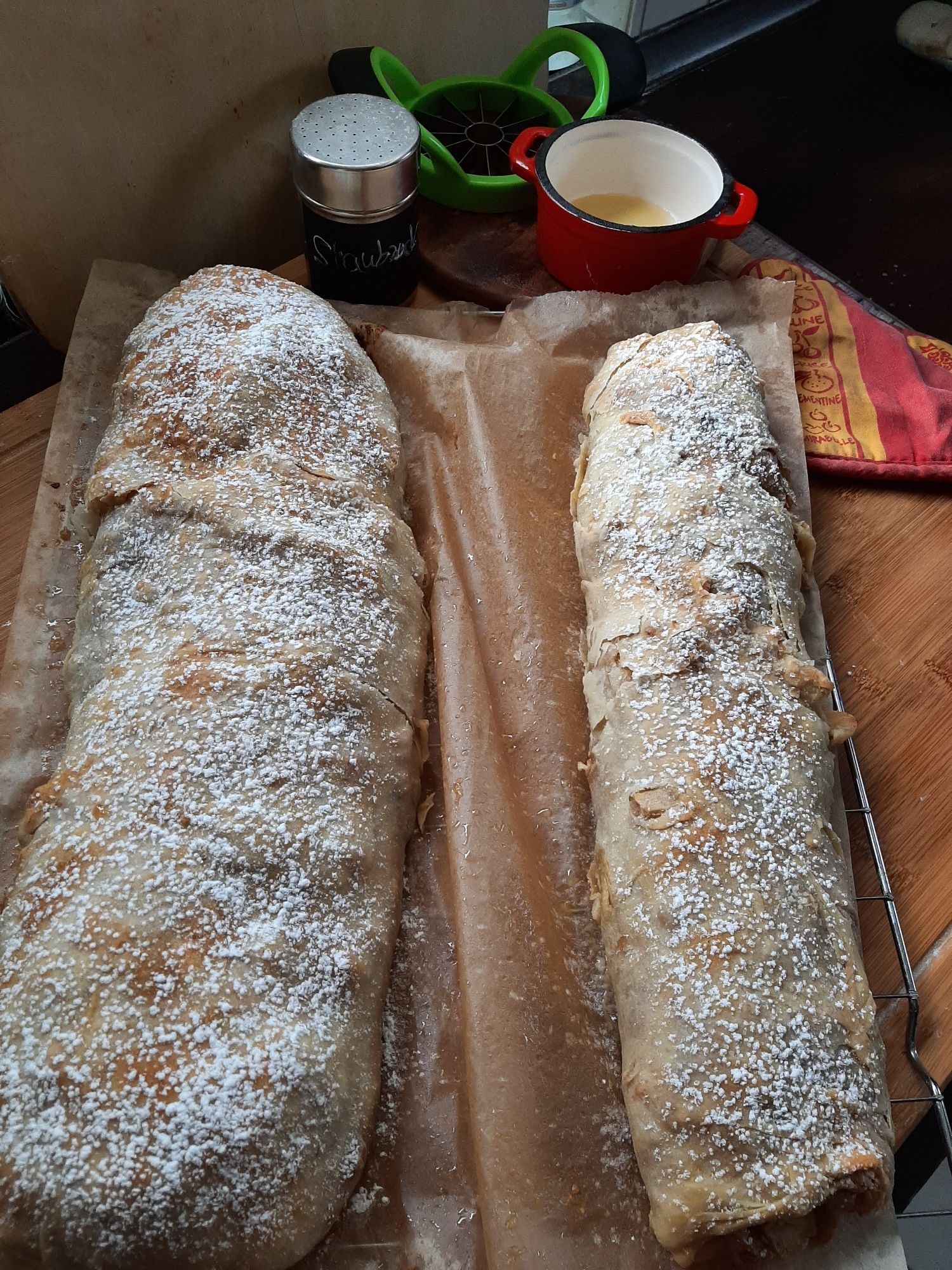 Zwei längliche Gebäckstücke. Sie sind mit Staubzucker bestreut. Im Hintergrund Backutensilien.