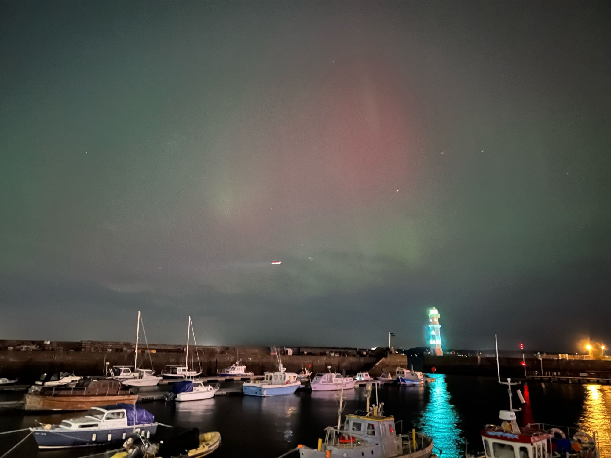 Looking towards a harbour, with boats and a lighthouse, the sky above has pink and green tinges from the Northern Lights.