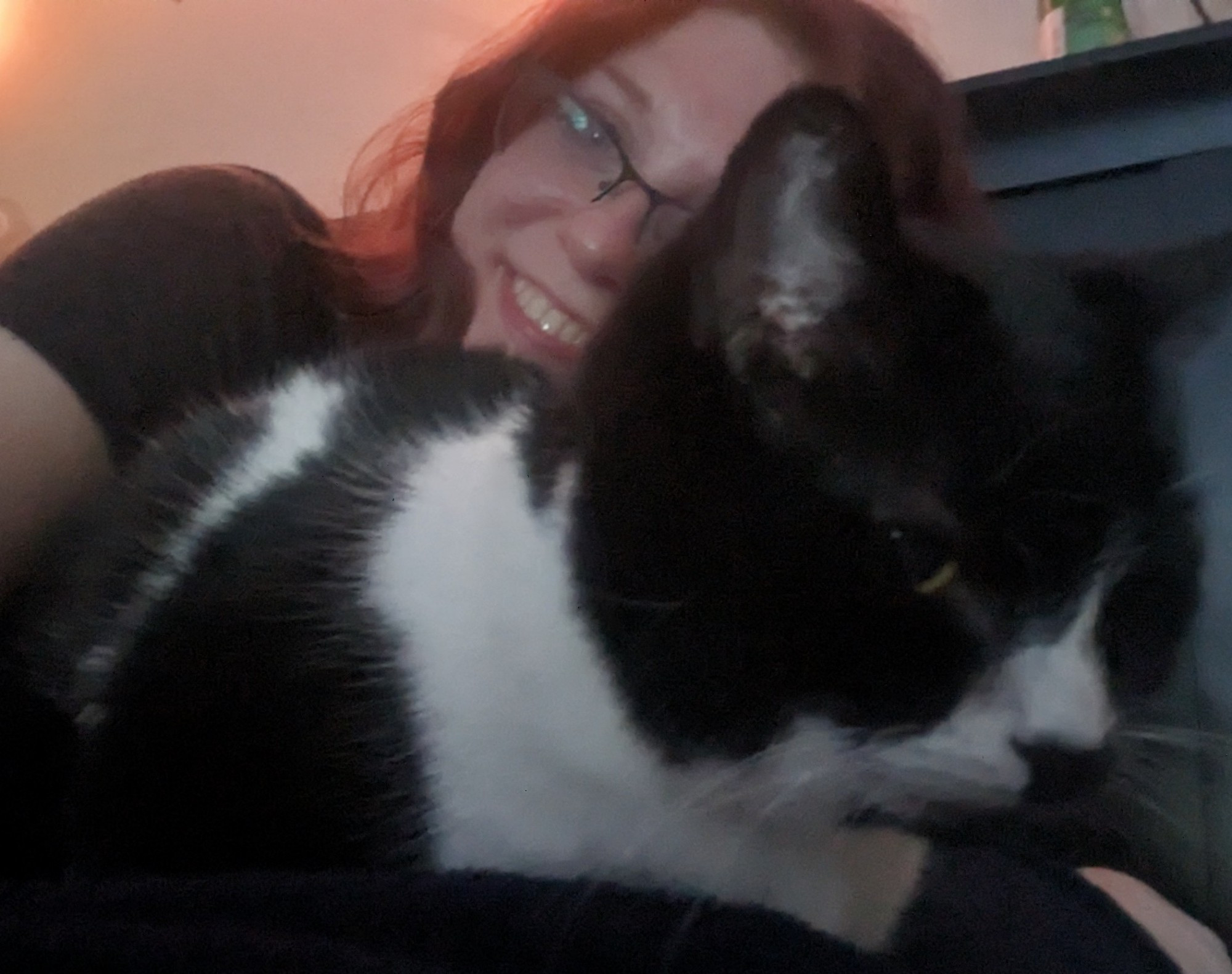 Effie laying in bed. Her tuxedo cat is curled up by her side in the fore front of the camera. 