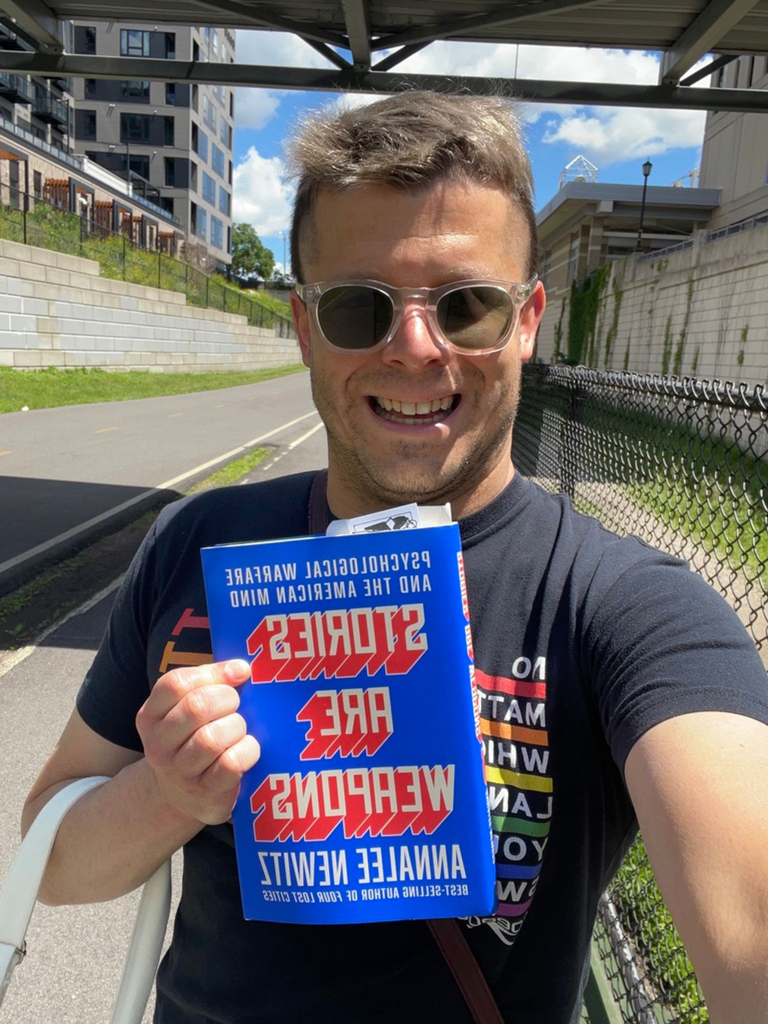 Me, in sunglasses + black swimming Pride shirt, holding STORIES ARE WEAPONS by Annalee Newitz; outside with blue sky/clouds on a bike/pedestrian path, smiling!