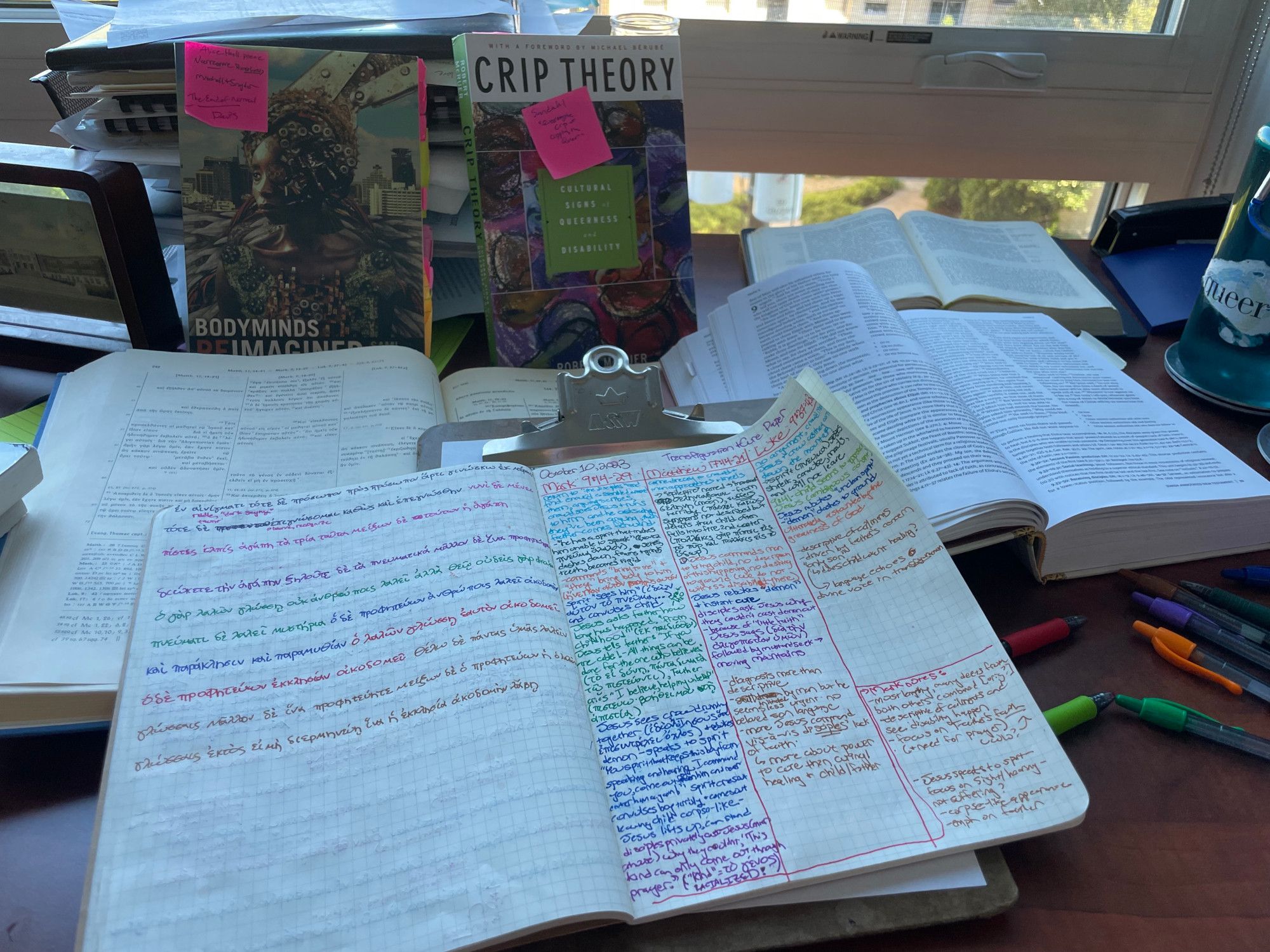 Image of a notebook filled with colorful notes paralleling three gospel texts, the notebook is on top of a full desk of research materials: a clipboard, a Bible and a Greek gospel synopsis and New Testament and two disability studies books (BODYMINDS REIMAGINED and CRIP THEORY)