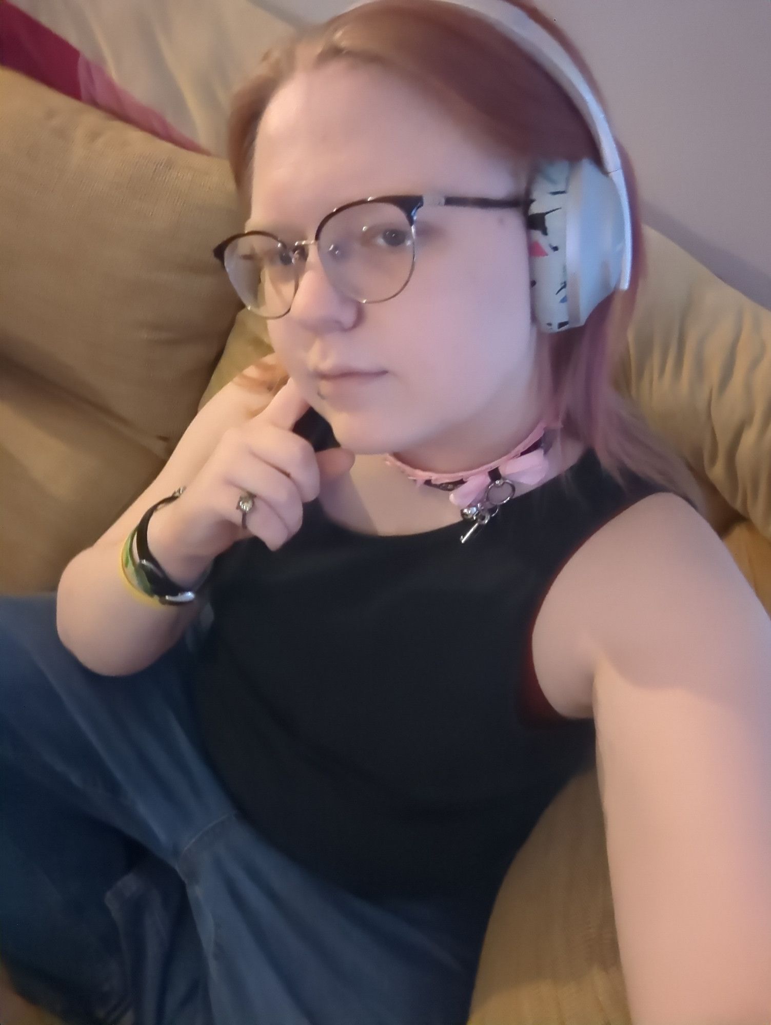 Many sitting on the couch in blue jeans and a black tank top, as well as headphones sweeping back faer hair and a pink and black lacy collar.