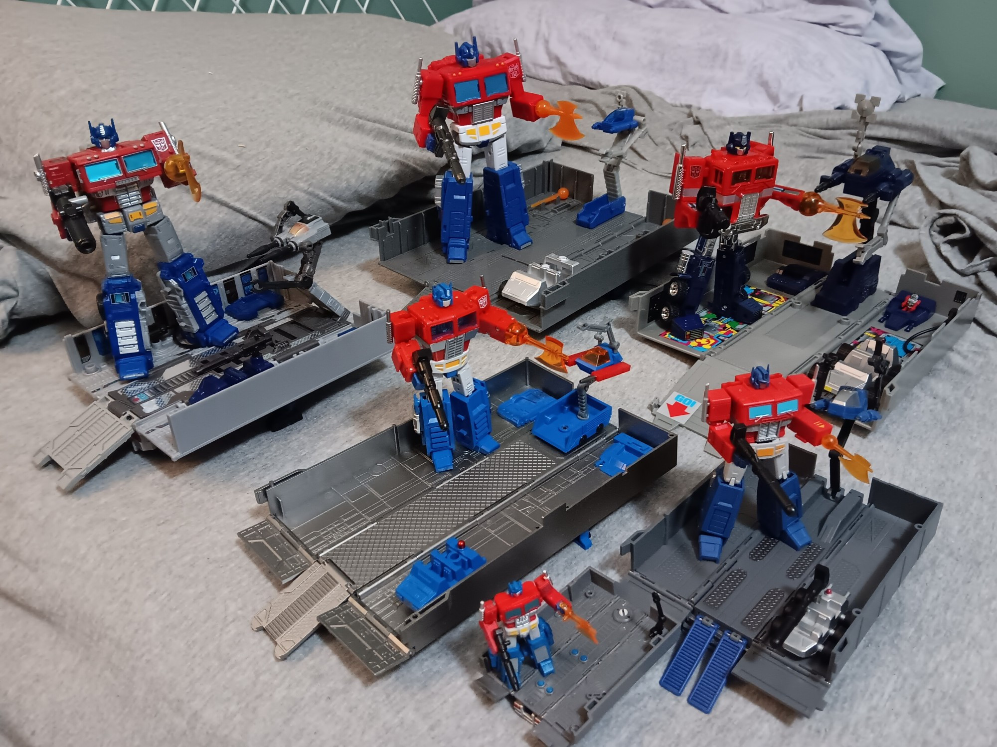 6 open combat deck trailers of various sizes set up on a bed, each with an Optimus Prime wielding his ion blaster and energon axe. Clockwise from furthest left,  Earthrise Leader, SS86 Commander, Missing Link Toy Deco, Magic Square Legends v1, Dr Wu , Hot Soldiers Prop Sky Pillar. Transformers toys