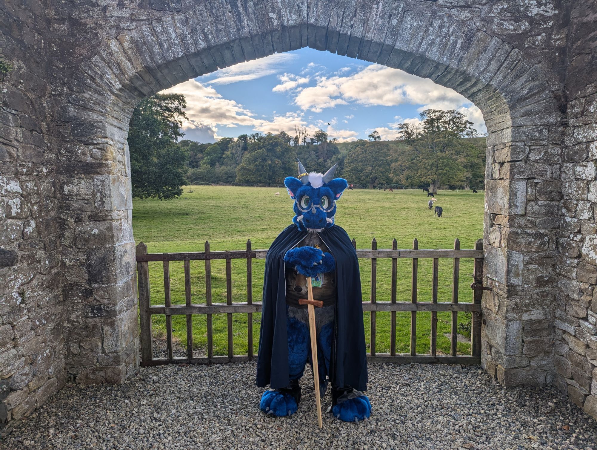 Blue dragon fursuiter Doran stands guard at a castle gateway with a green field behind him, wearing a blue cloak and resting his handpaws on the hilt of a wooden longsword.